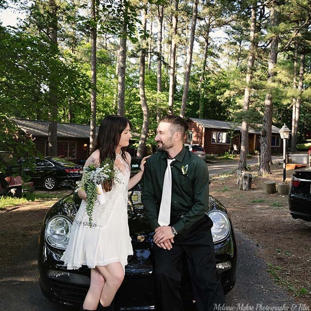 A couple smiles at each other while sitting on a black car in a forested setting.
