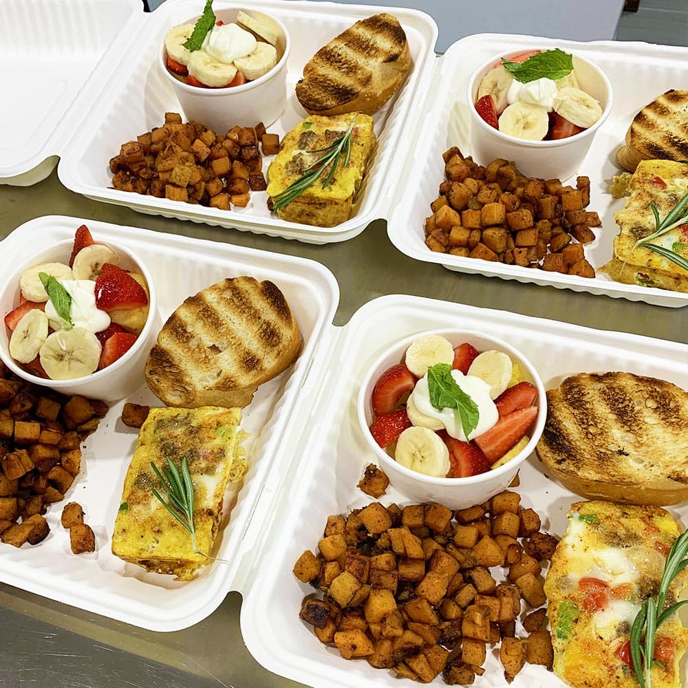 Four meal trays with egg frittatas, roasted potatoes, grilled bread, and fruit bowls of strawberries and bananas.