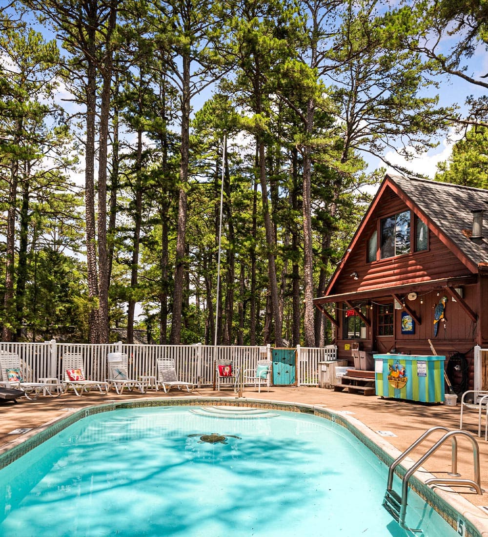 A swimming pool surrounded by tall trees and a cabin with outdoor seating.