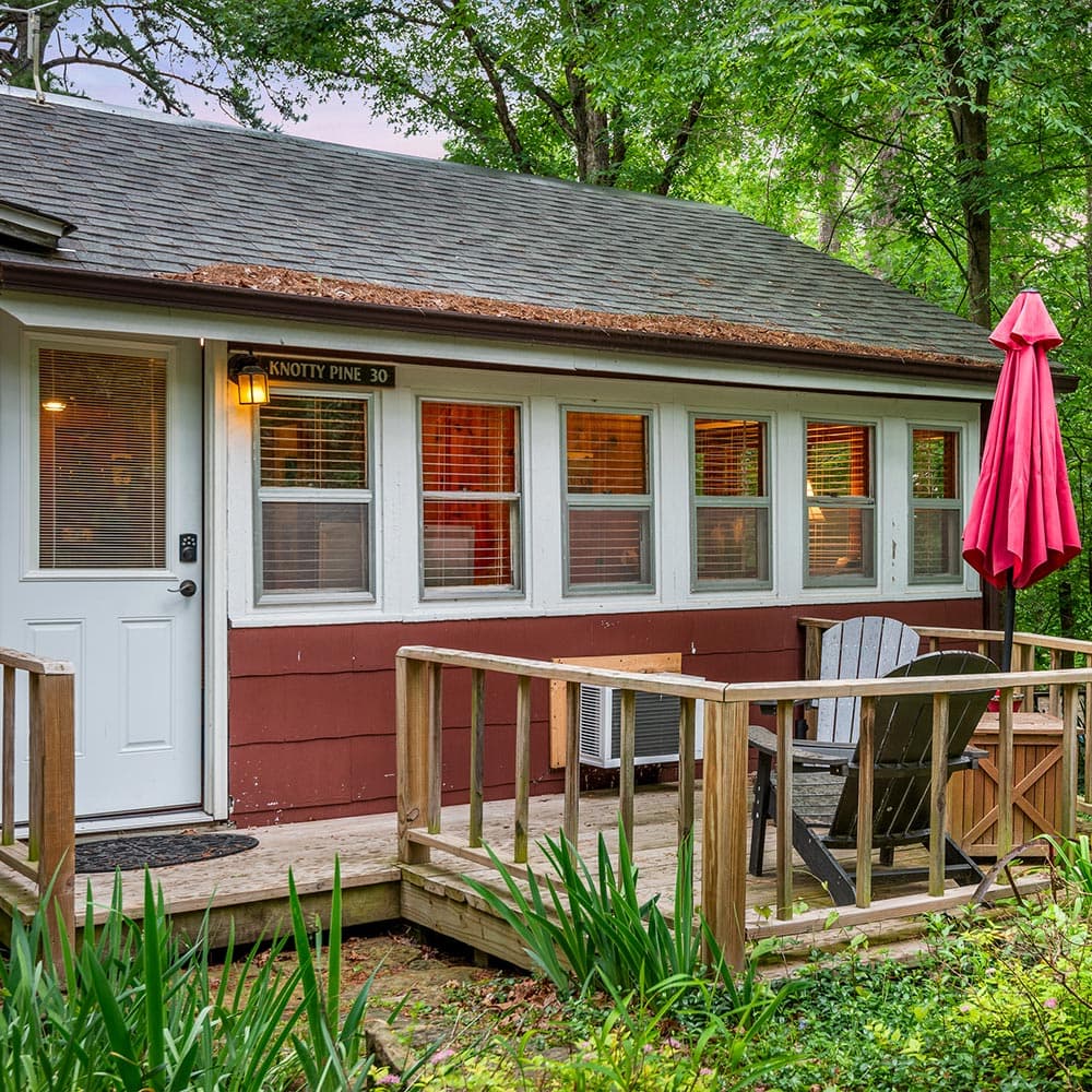 A cozy cabin with a wooden deck and a red umbrella, surrounded by greenery.