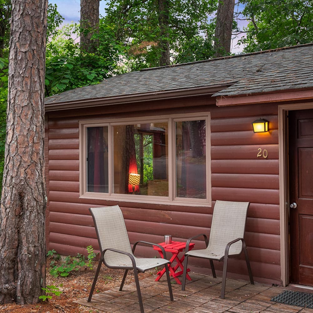 A cozy cabin surrounded by trees, featuring a small table and chairs on a stone patio.