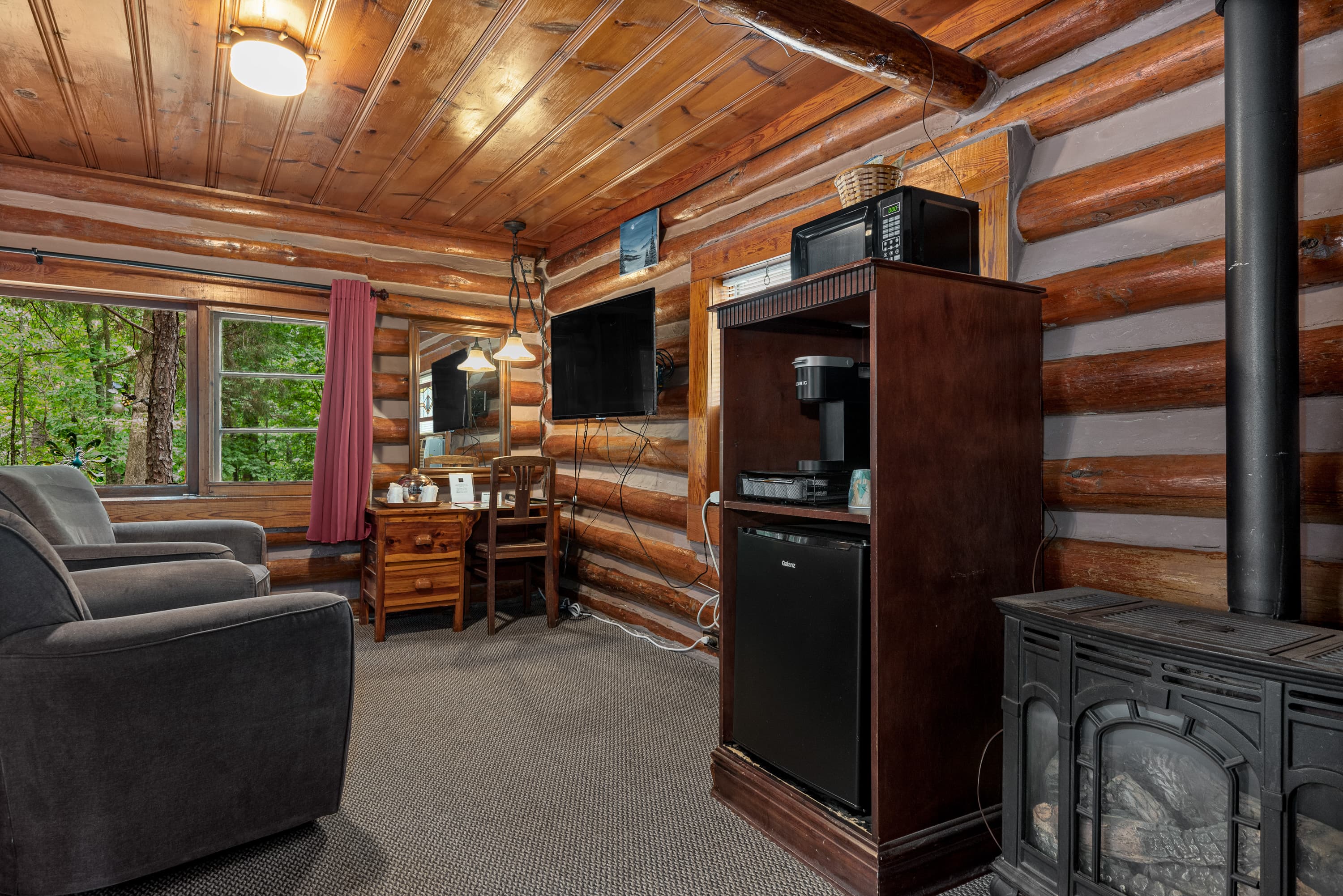 A log cabin bedroom with a rustic wood stove, two armchairs, a TV, and a beverage center with a mini-fridge, microwave, and coffee maker.