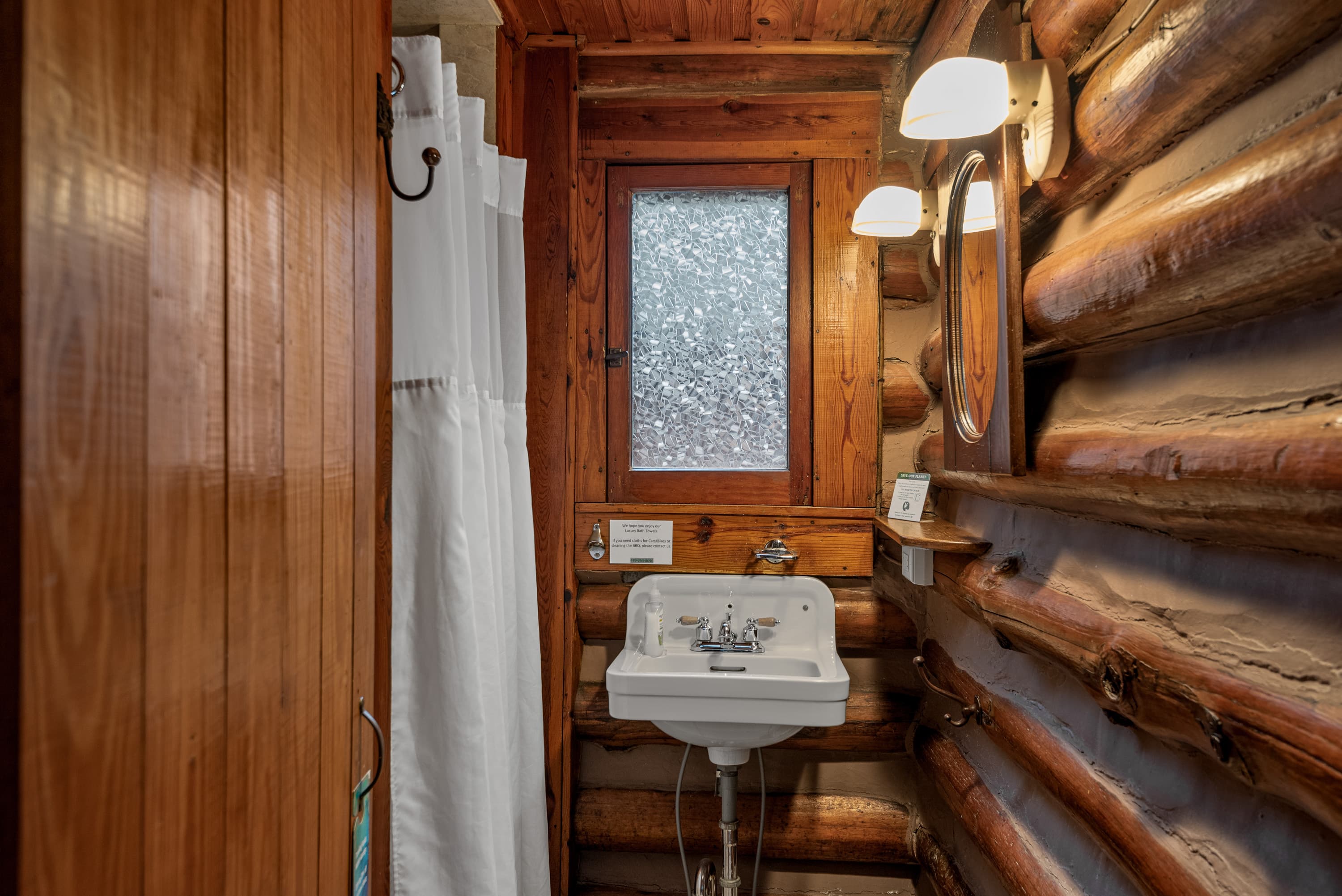 A cozy, rustic bathroom with a small, traditional sink, a mirror, and a window. The walls are made of natural wood logs.