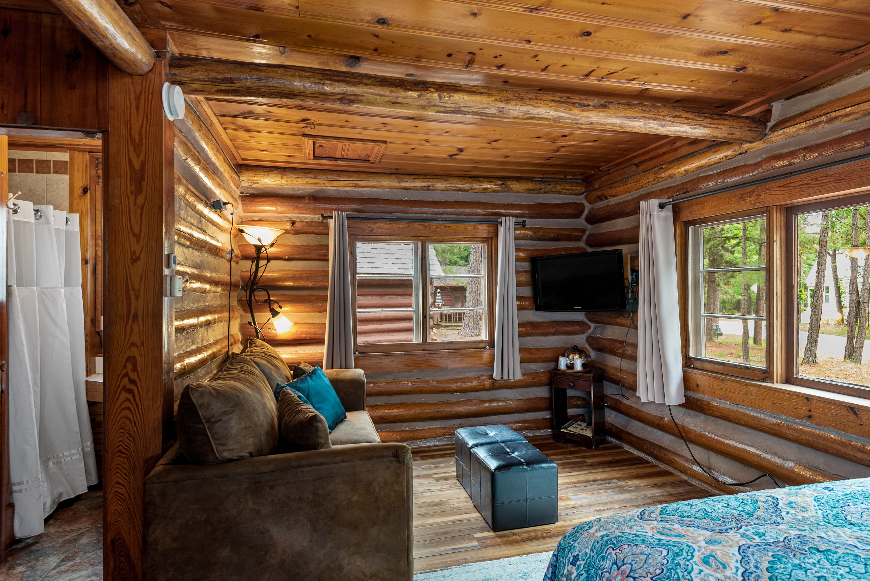 A small, cozy log cabin living room with a brown couch, a black ottoman, and a flat-screen TV. The rustic walls are made of logs.