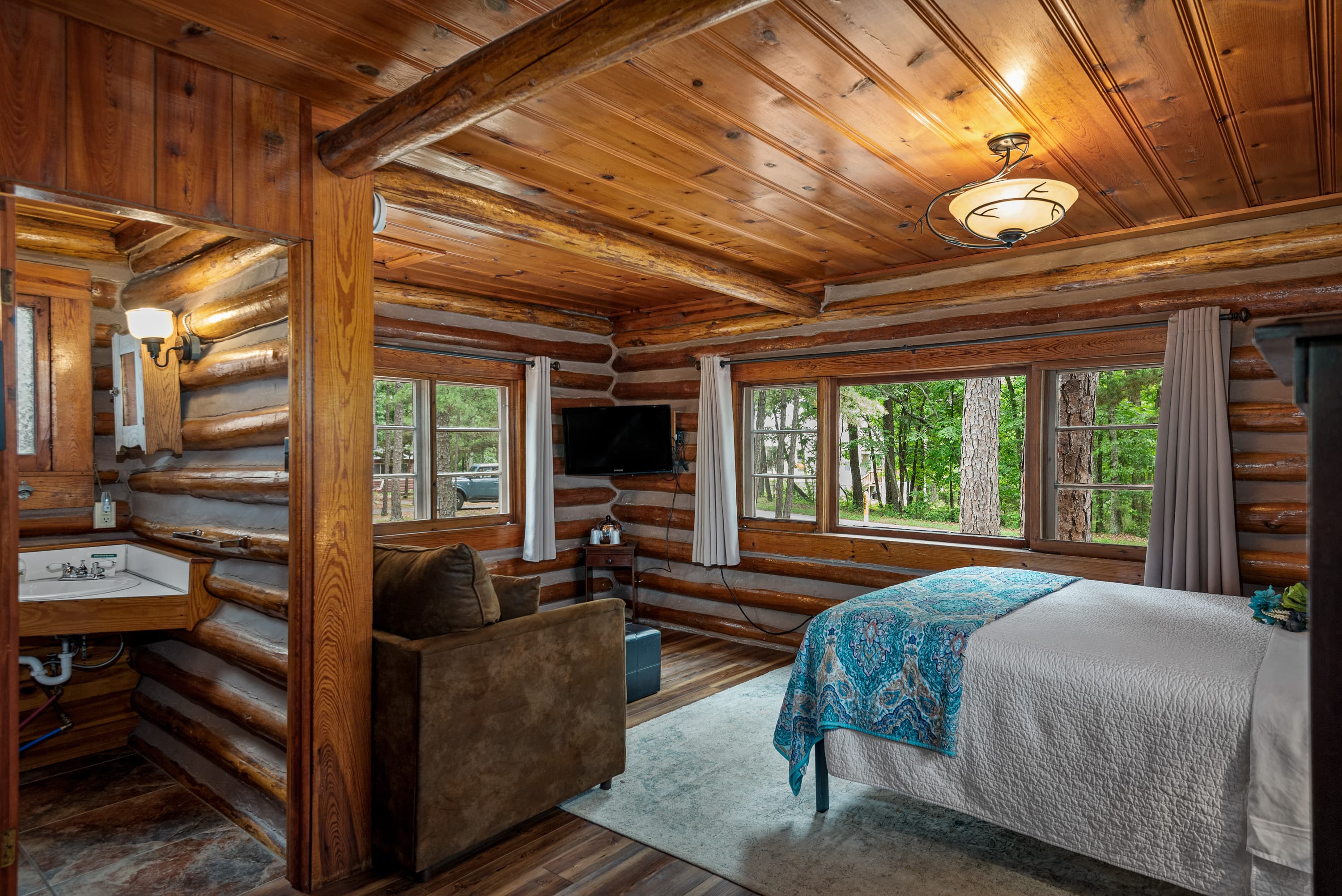 A small log cabin studio with a bed, a TV, a couch, and a rustic sink area. The walls, ceiling, and floor are made of natural wood.
