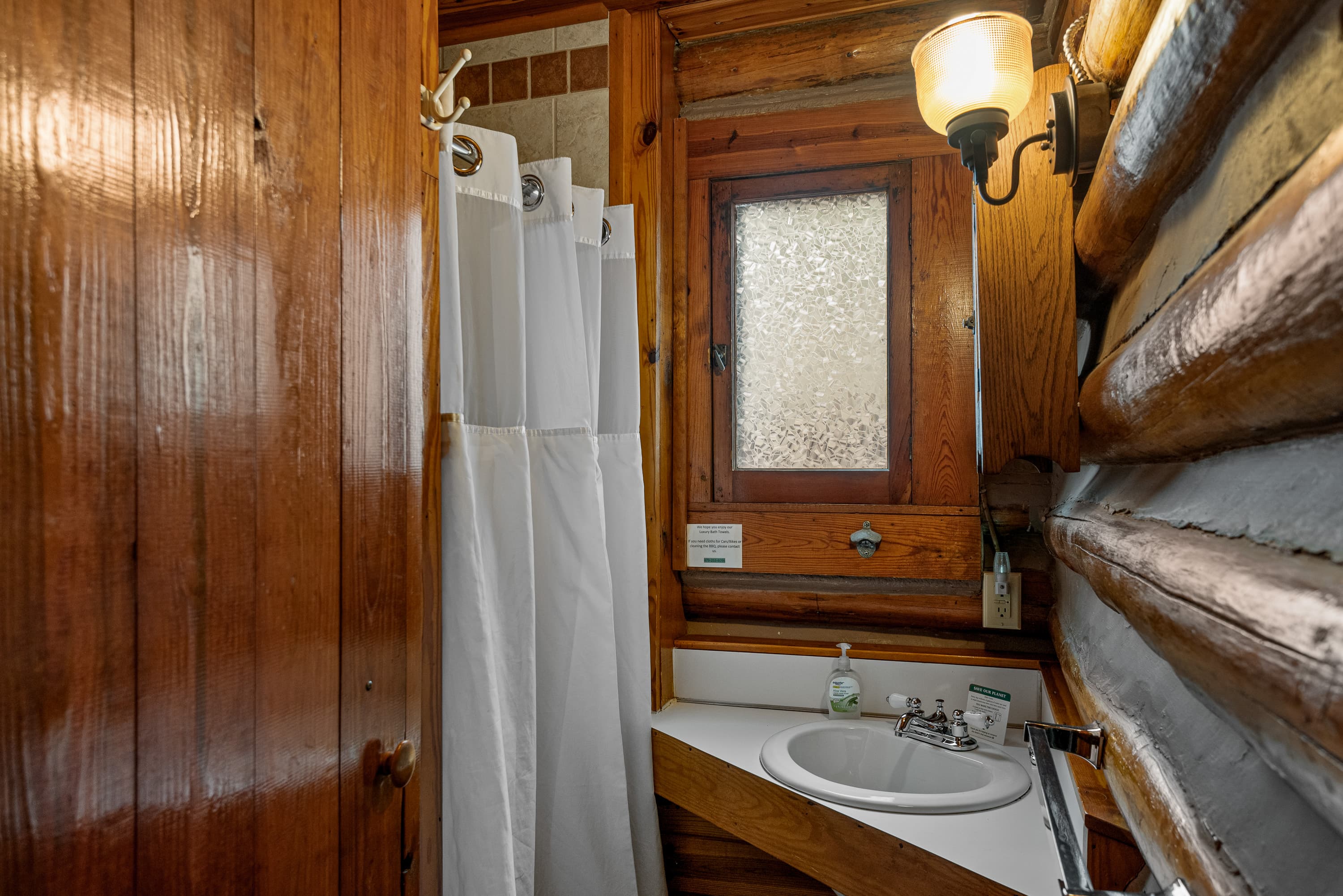 A compact log cabin bathroom with a white shower curtain, a small sink with a mirror, and a window. The walls are a mix of wood planks and logs.