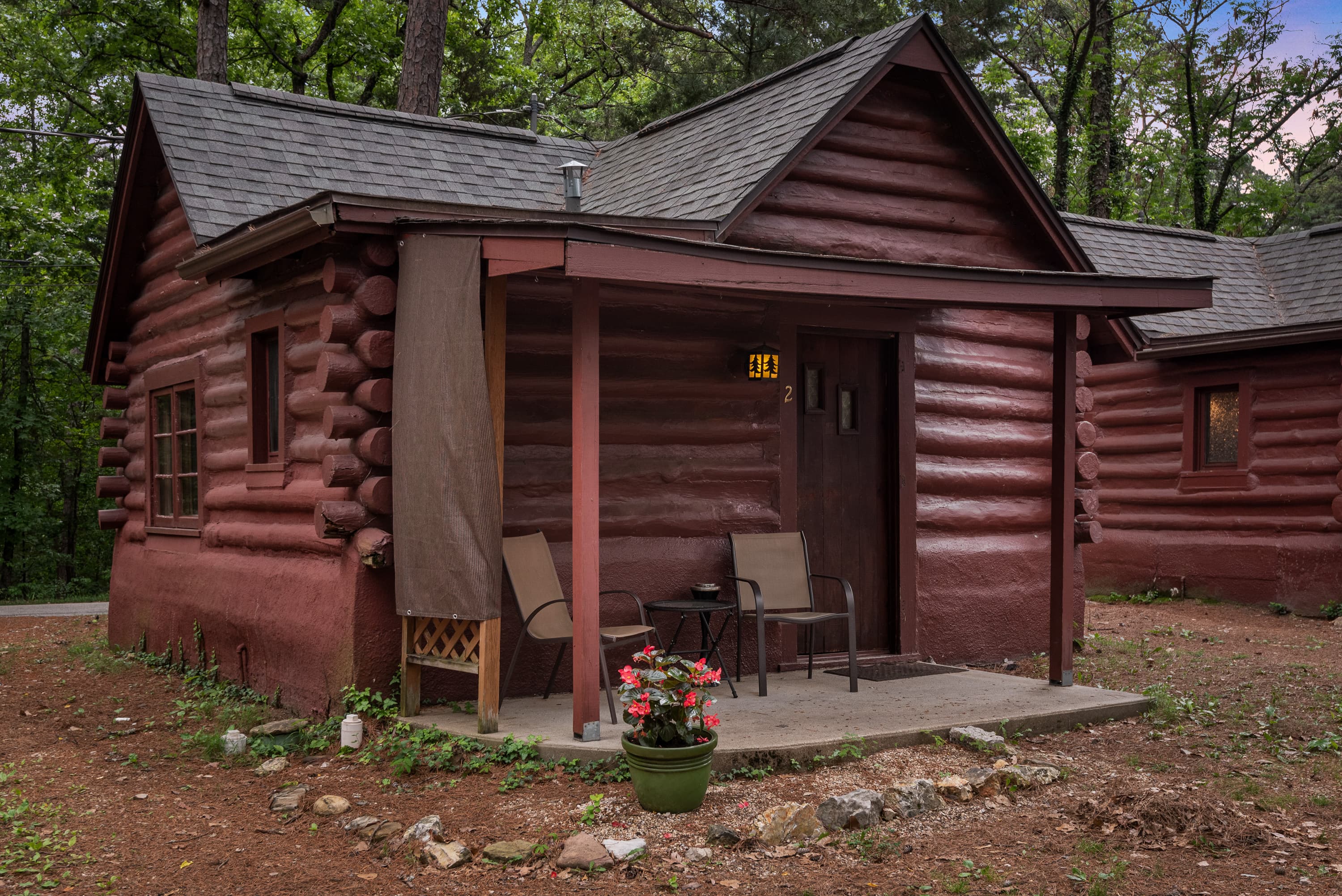 A cozy, small log cabin with a covered porch holding two chairs. The cabin is surrounded by a small garden with a trellis and lush greenery.