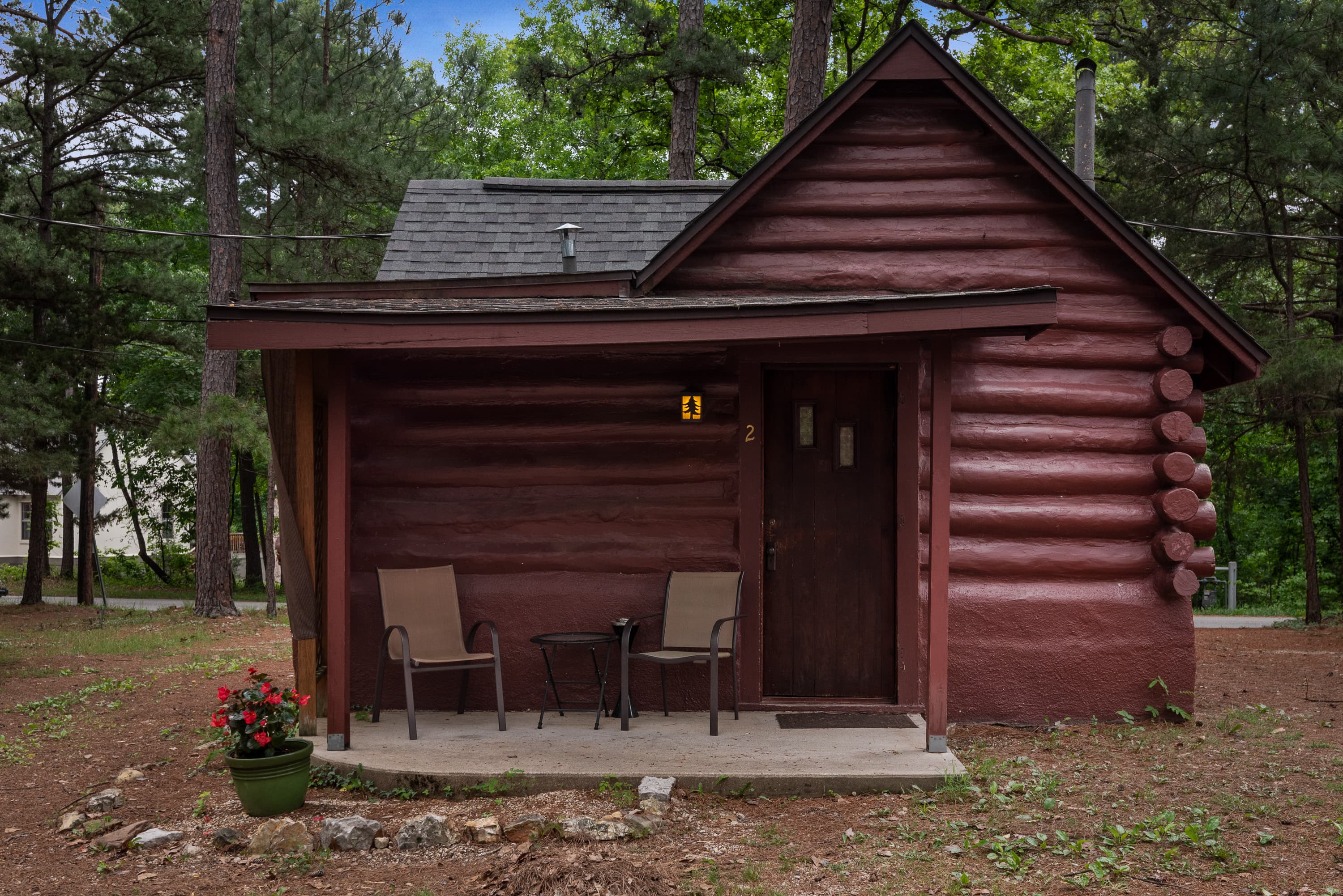 A rustic log cabin with a covered porch holding two chairs and a small table. A pot of red flowers sits on the ground nearby.