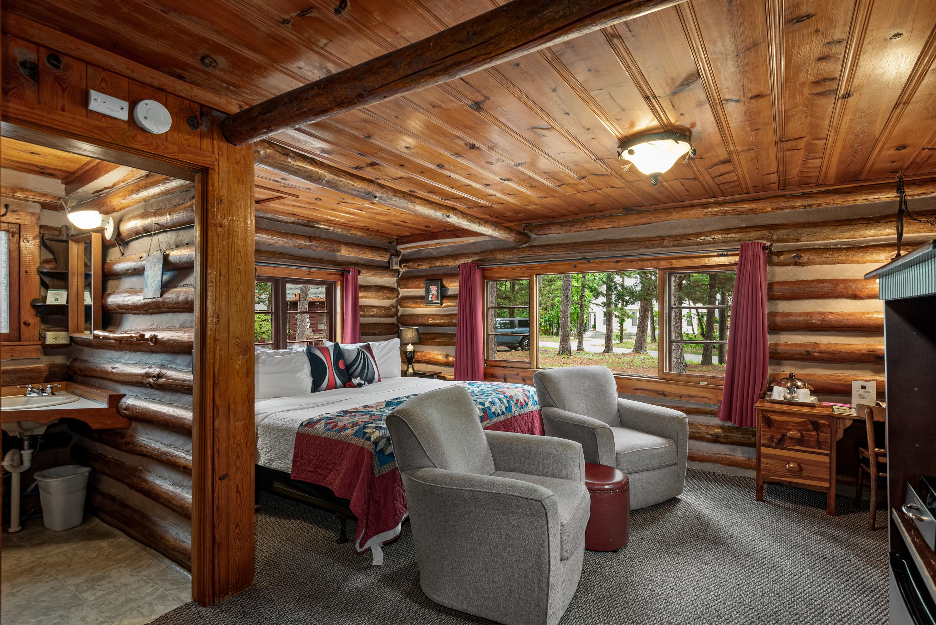 A spacious log cabin bedroom with a king bed, two armchairs, a small desk, and a rustic sink area. The walls are made of natural wood logs.