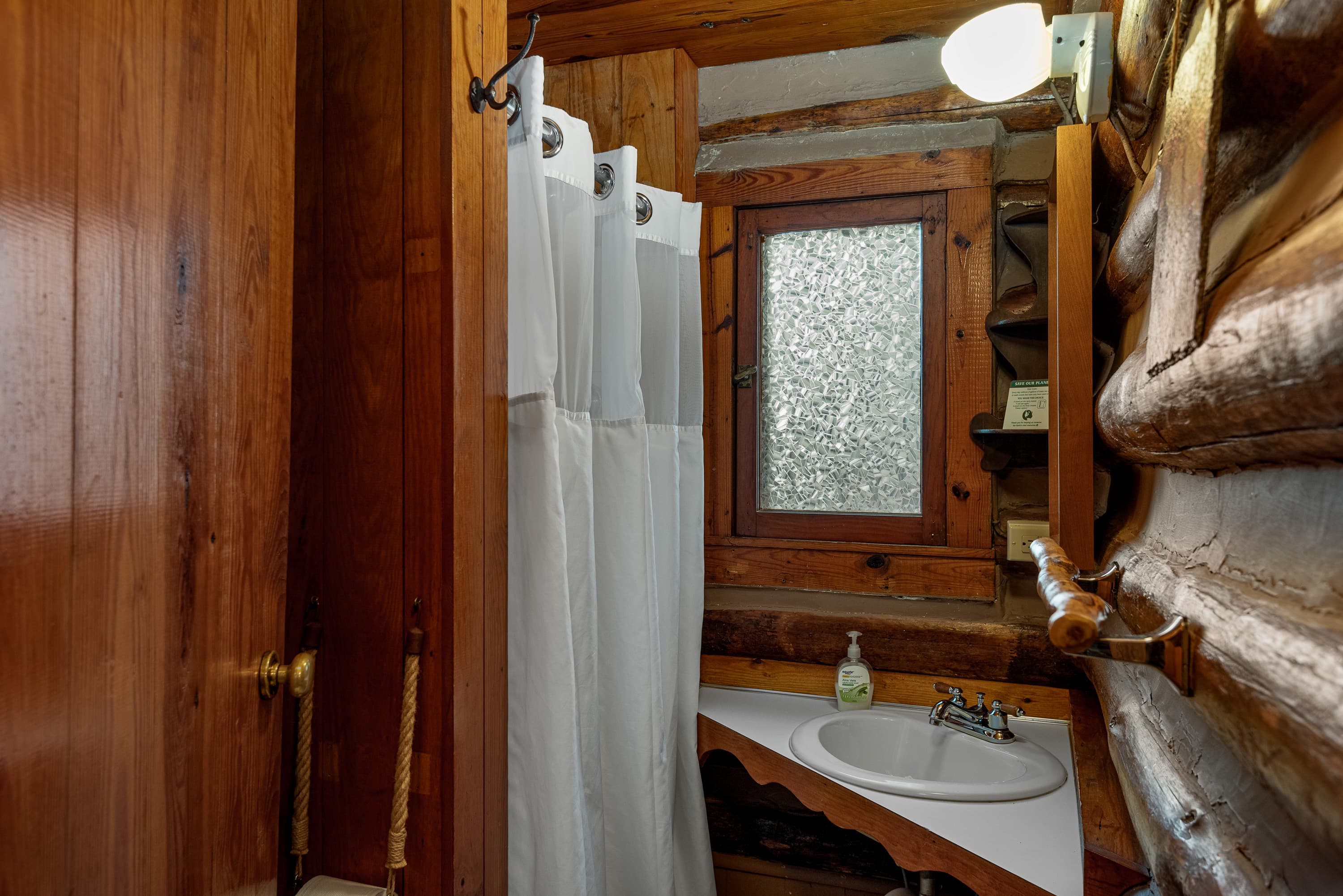 A small log cabin bathroom with a white shower curtain, a corner sink with a wood counter, and a window.