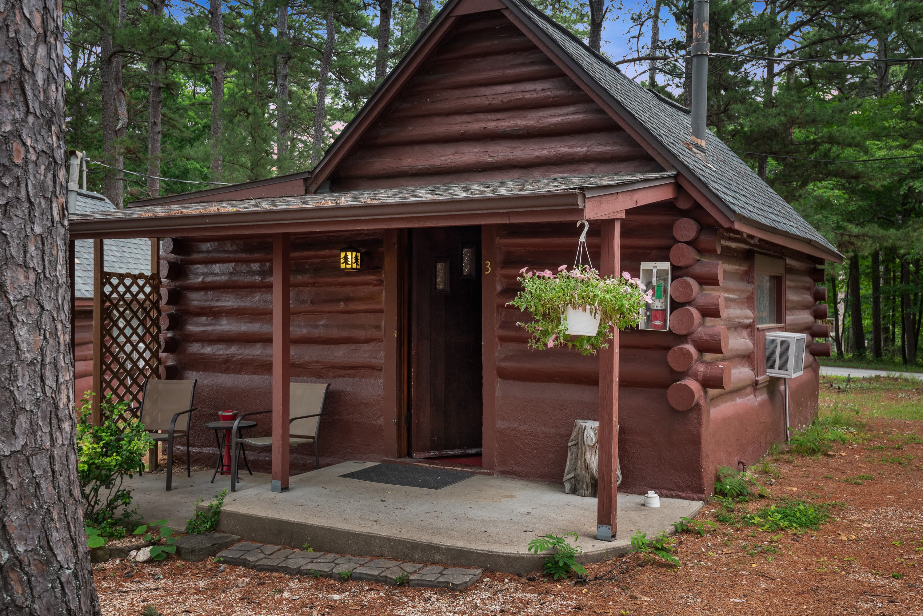 A charming, small log cabin with a covered porch holding two chairs. The cabin is surrounded by a small garden with a trellis and lush greenery.