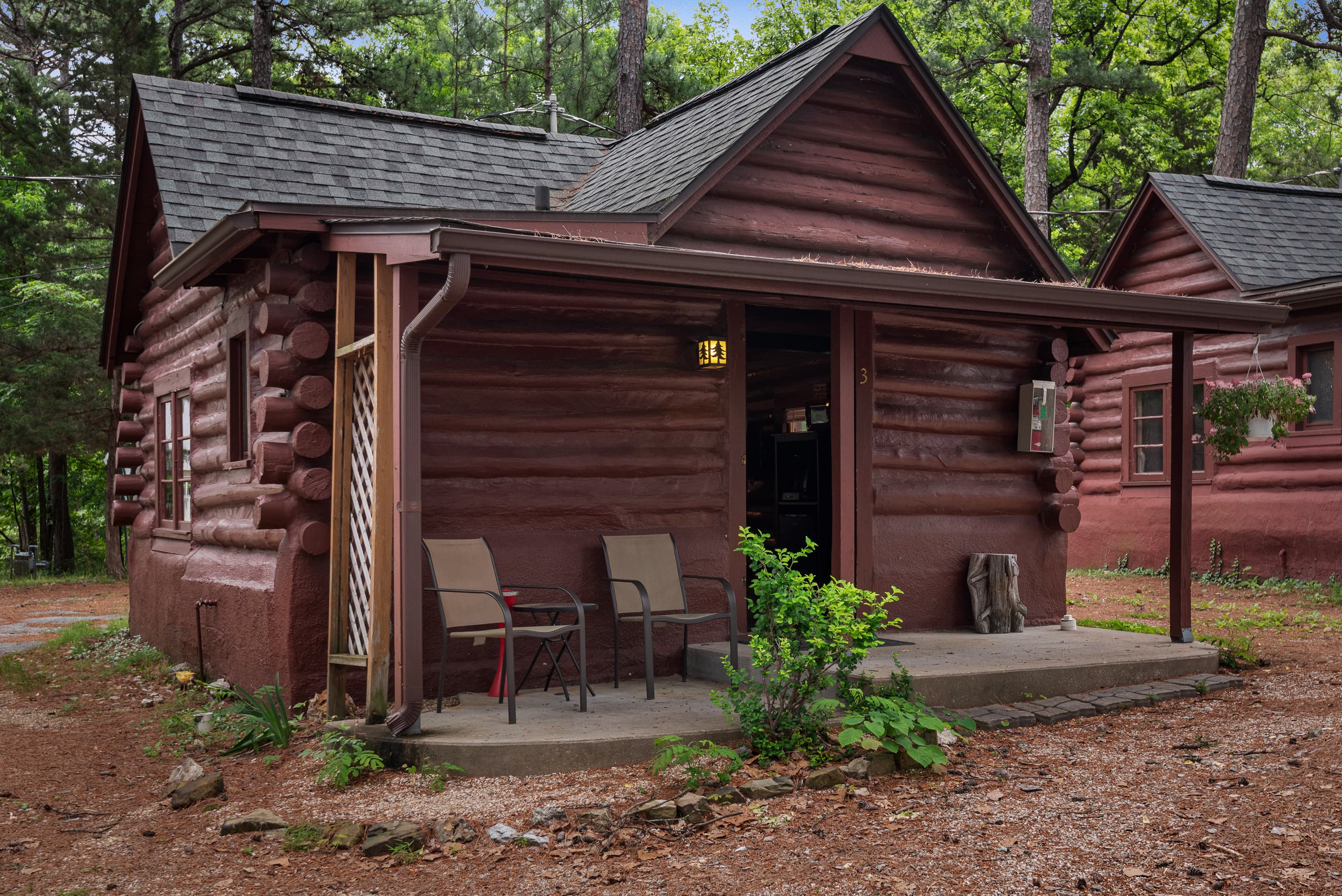 A small, rustic log cabin with a covered porch holding two chairs. The cabin is surrounded by a natural wooded area with pine needles on the ground.
