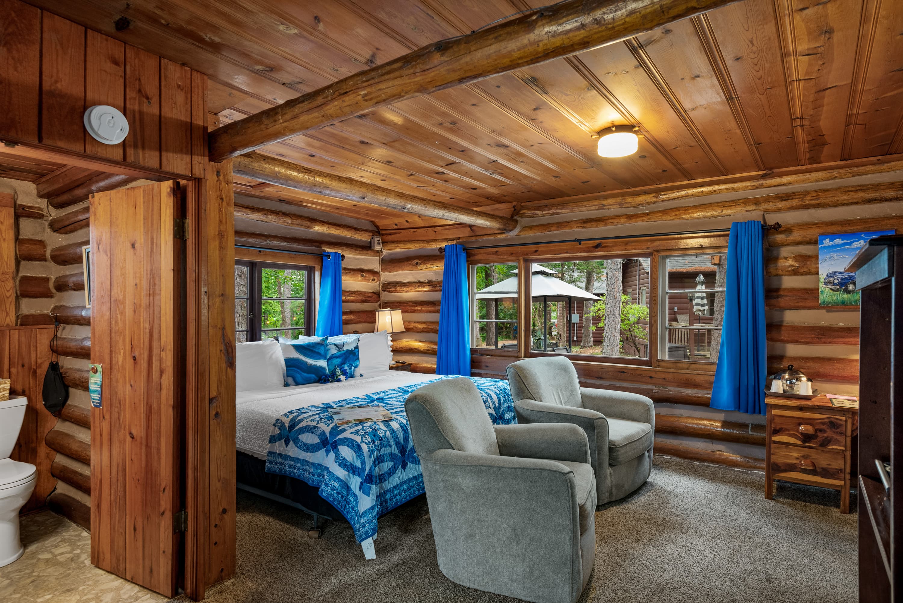 A log cabin bedroom with a king-size bed featuring a blue quilt. The room includes two armchairs, a television, and rustic wood-log walls.