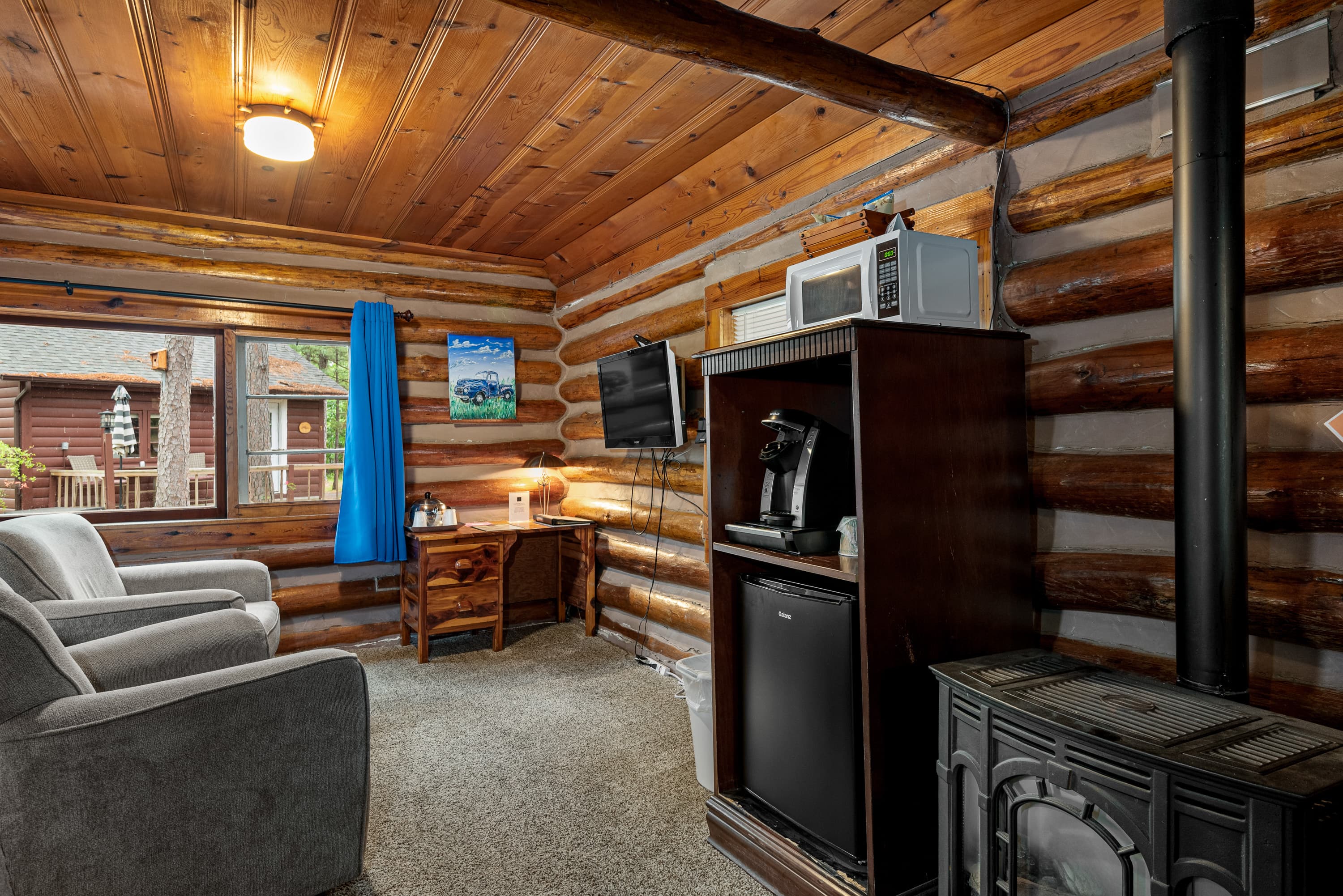 A cozy log cabin room features two armchairs, a television, a small desk, and a rustic wood stove.