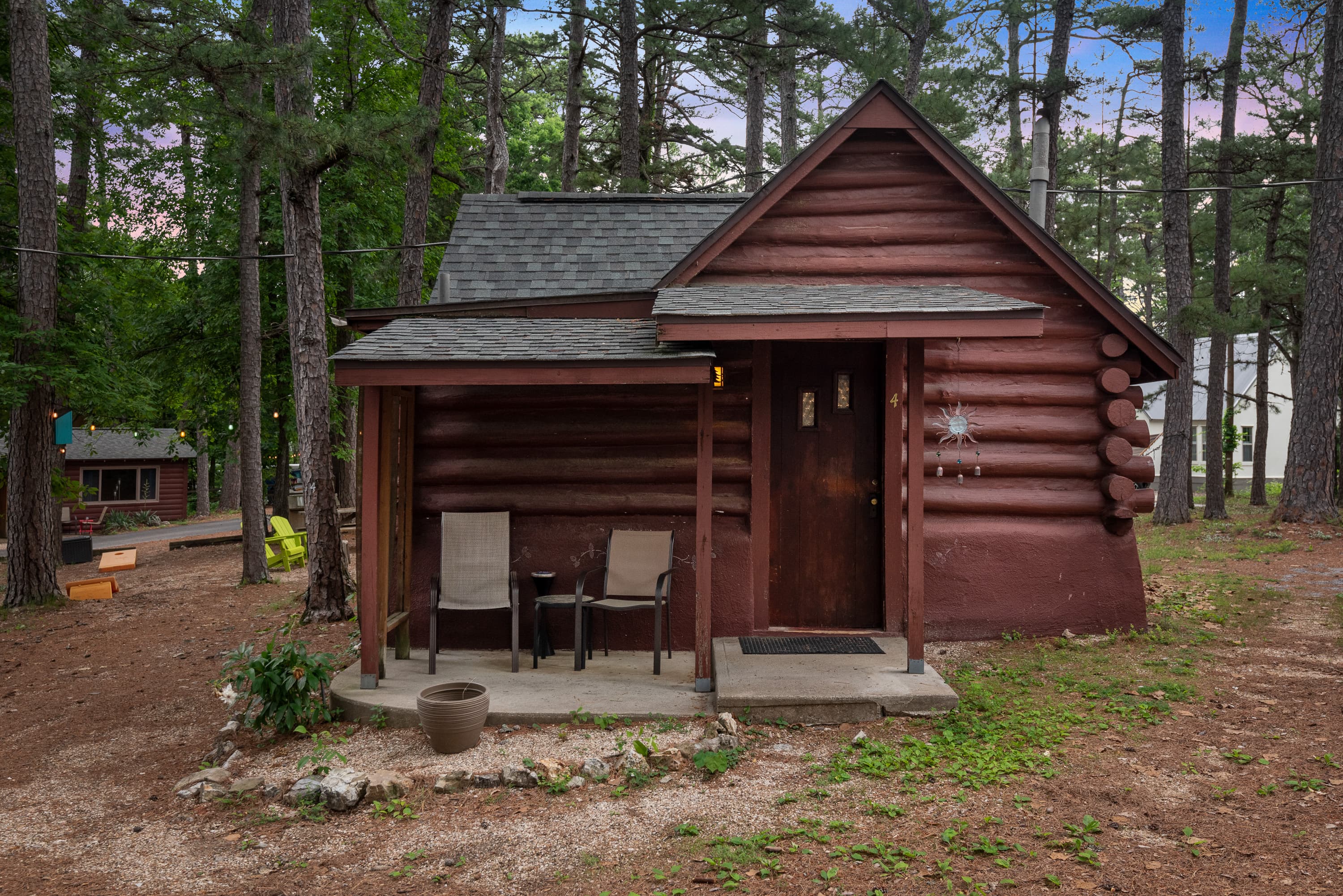 A cozy, rustic log cabin with a covered porch holding two chairs. The cabin is surrounded by a natural wooded area with pine trees.