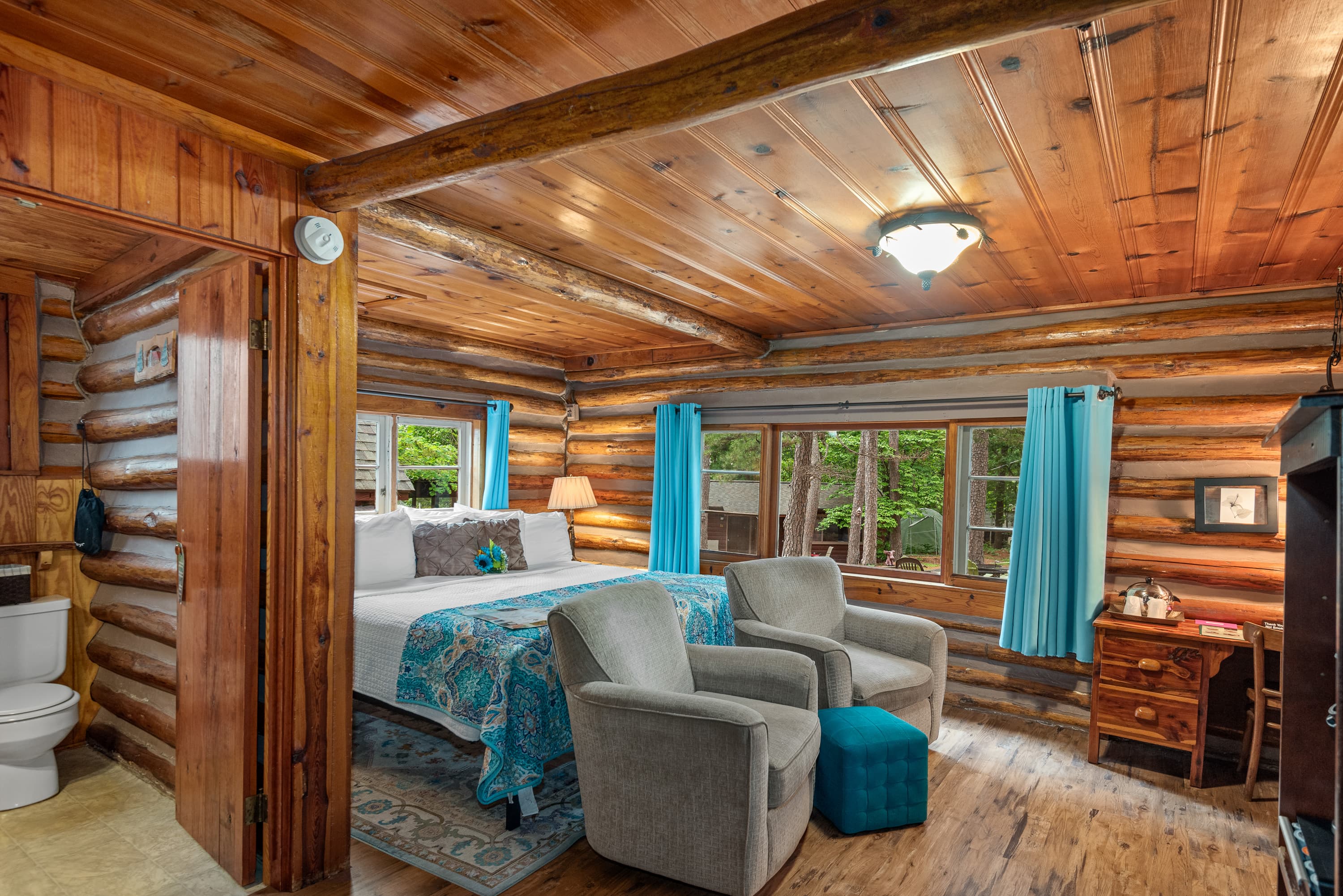A spacious log cabin bedroom with a king bed, two armchairs, and a small desk.