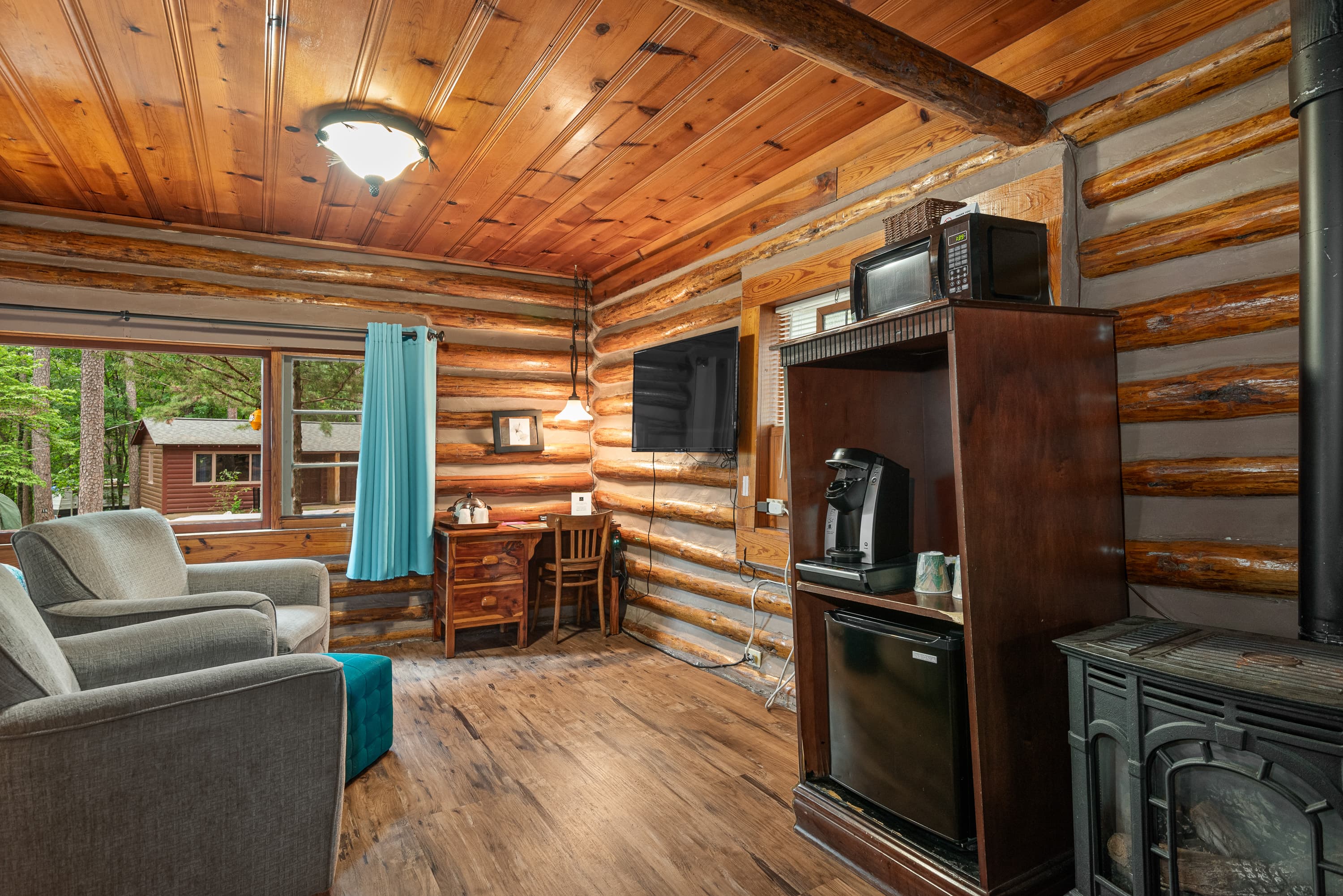 A cozy log cabin room features two armchairs, a television, a small desk, and a rustic wood stove.