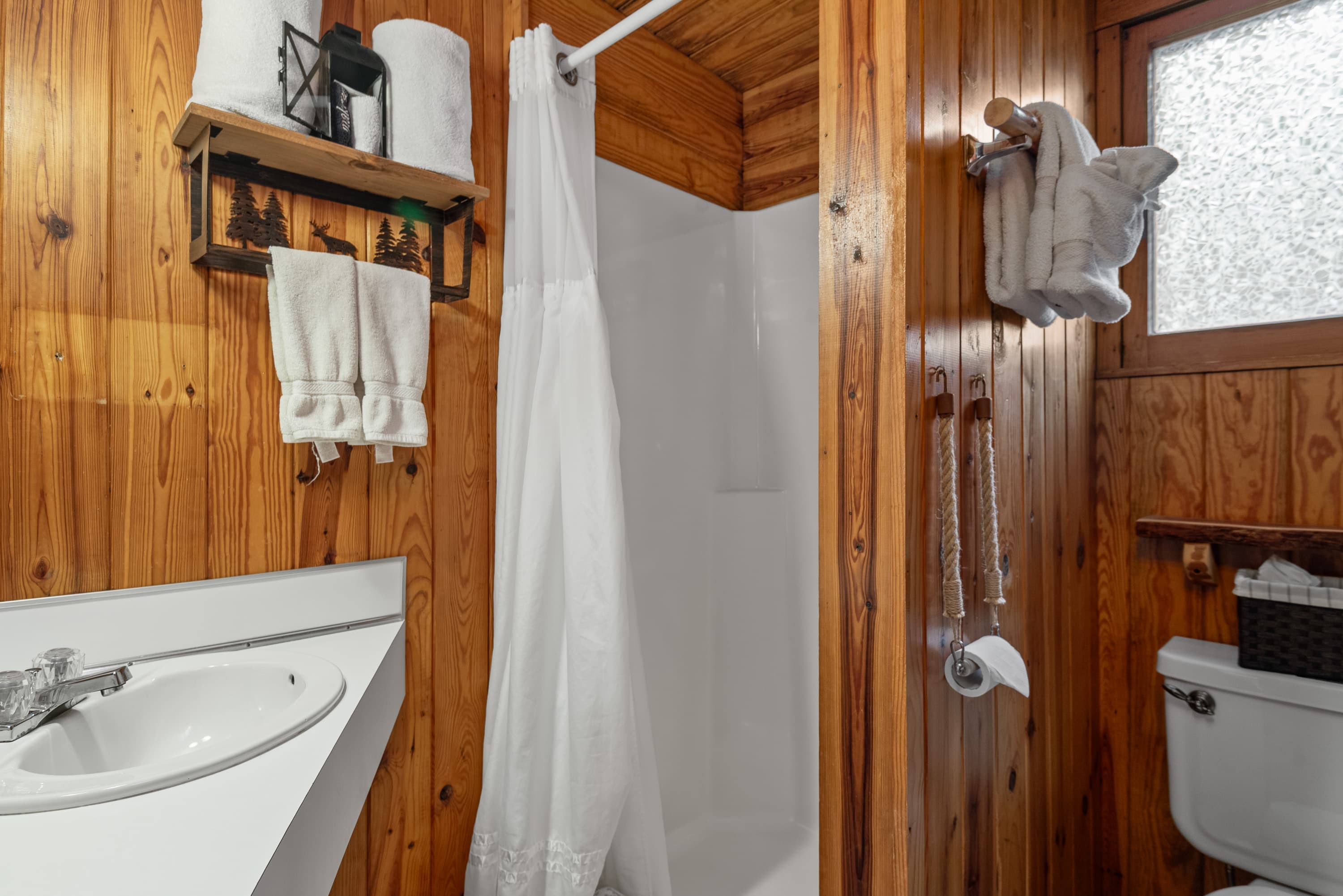 A cozy, compact bathroom with wood-plank walls, a stand-up shower, a white sink, a toilet, and a white shower curtain. Towels are stacked on a shelf.