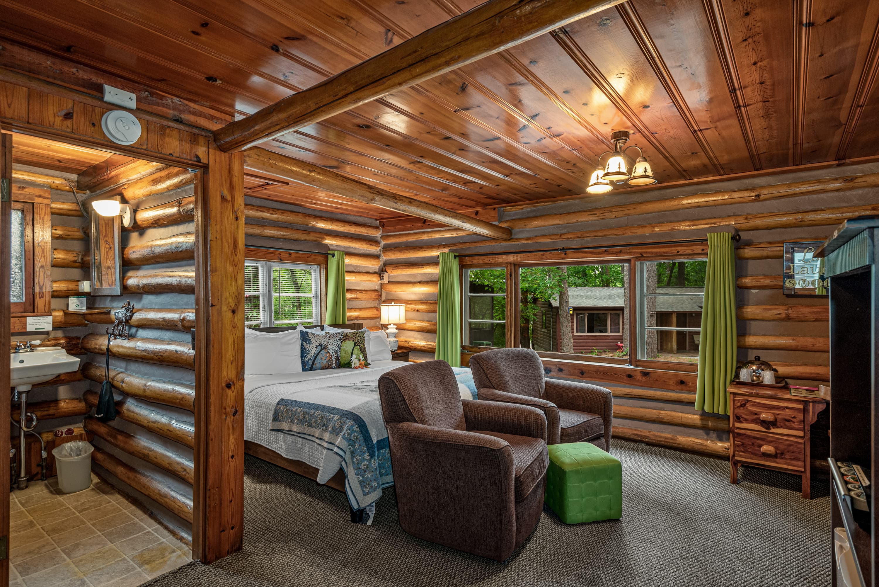A log cabin interior with a bed, two armchairs, a green ottoman, and a rustic sink area.