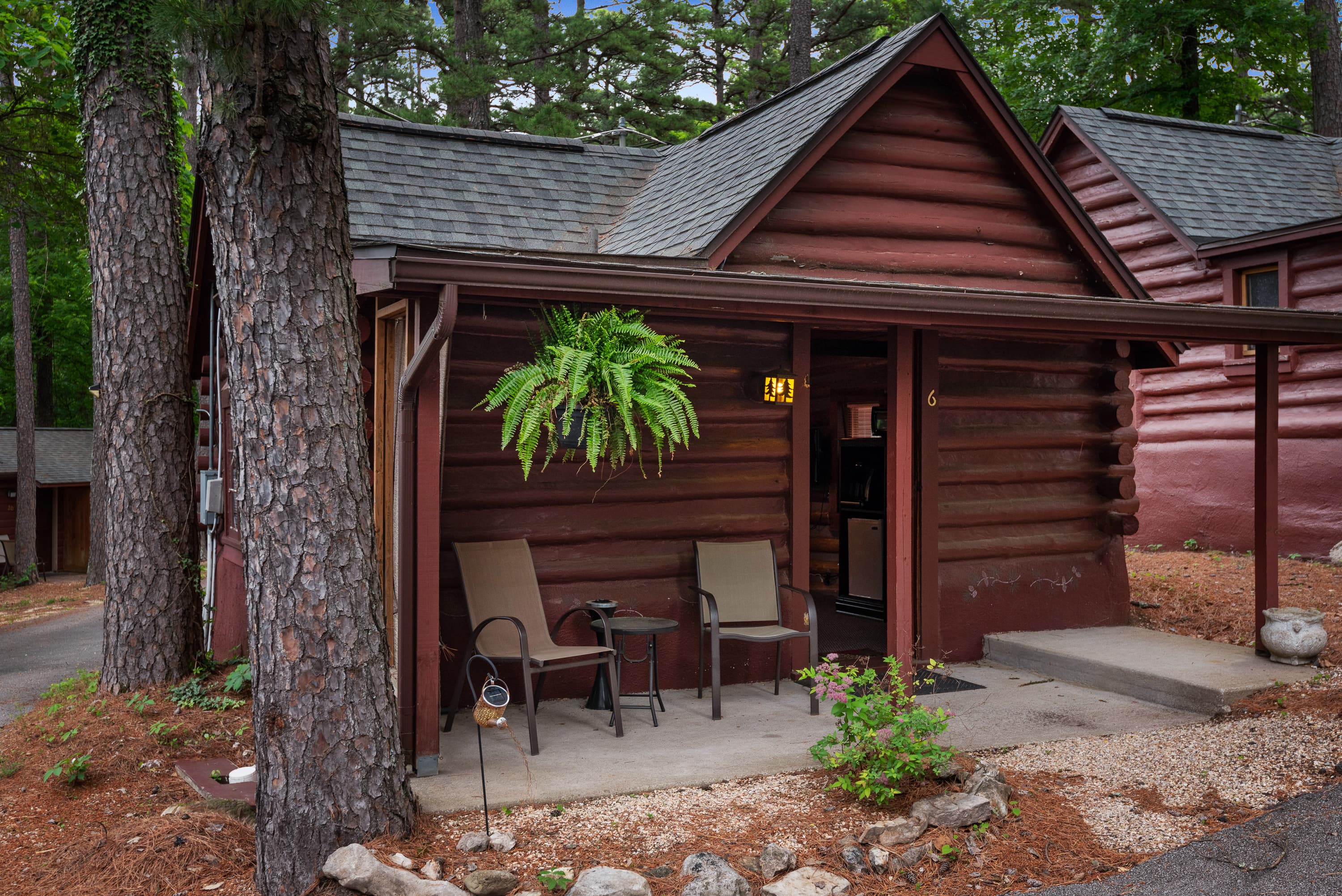 A charming log cabin is nestled among trees, with a covered porch holding two chairs and a small table. A fern in a hanging basket hangs on the porch.