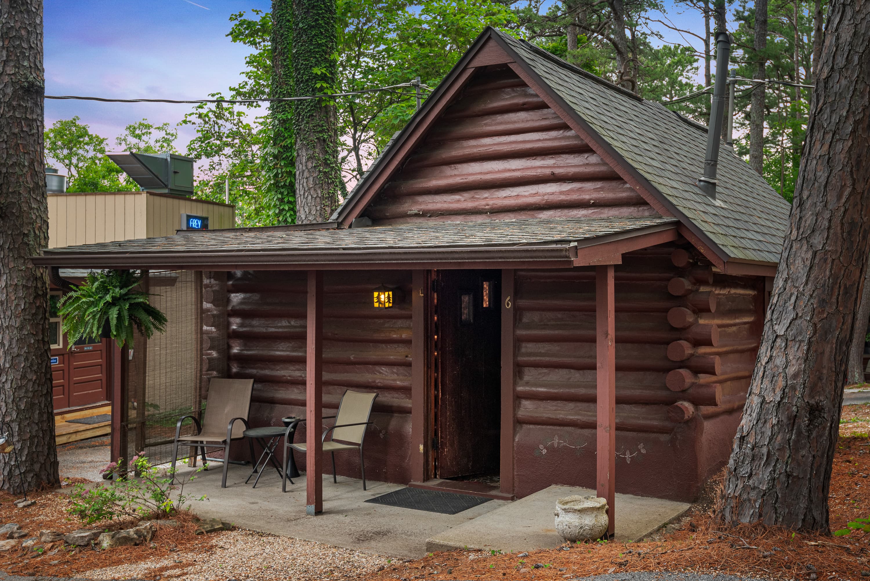 A cozy, small log cabin with a covered porch holding two chairs. The cabin is surrounded by a natural wooded area with pine needles on the ground.