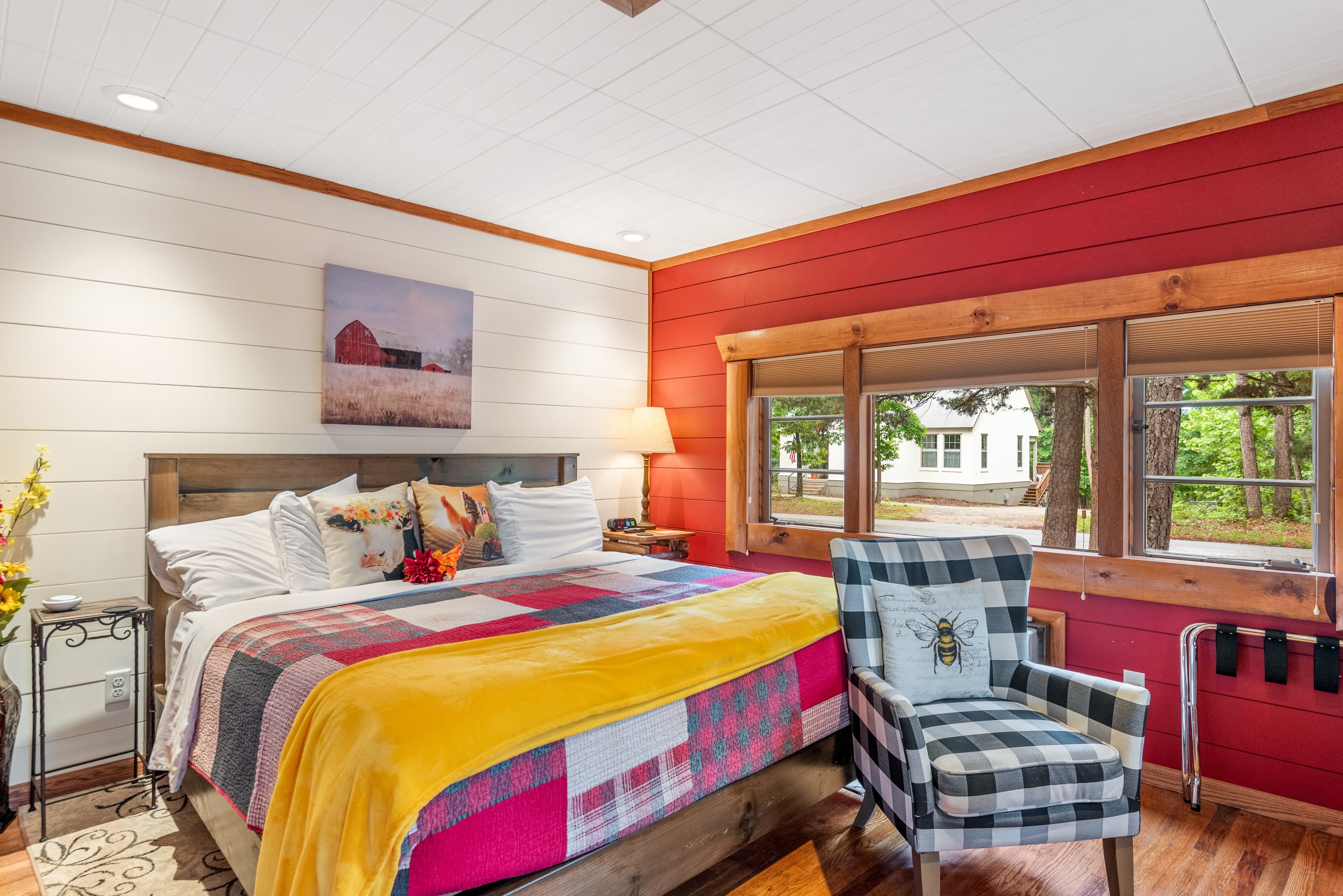 A rustic bedroom features a king-size bed with a colorful patchwork quilt. The room has white and red wood-plank walls and a black and white plaid armchair.