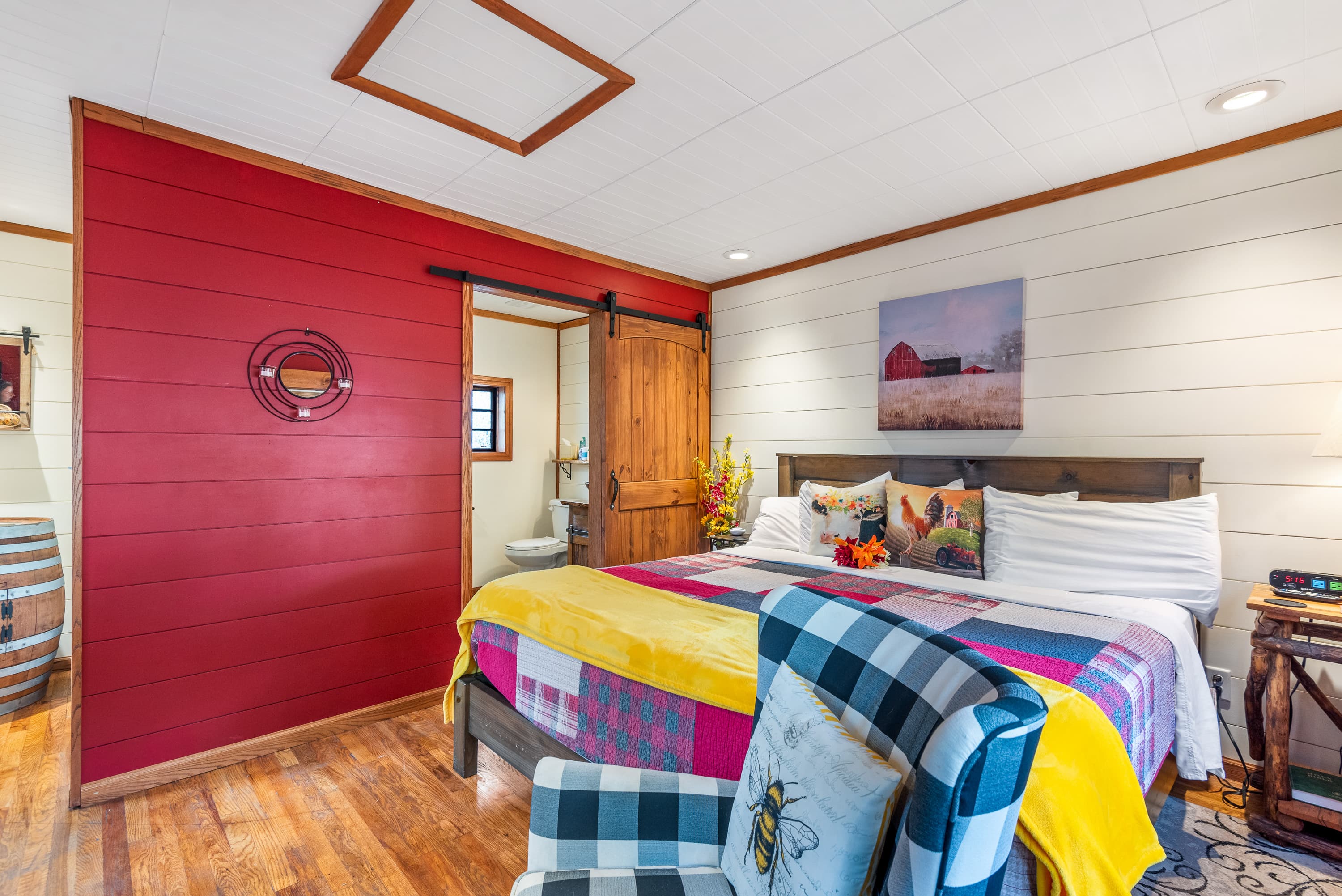 A cozy, rustic bedroom with a king-size bed, a patchwork quilt, and a red accent wall. A sliding barn door leads to the bathroom.