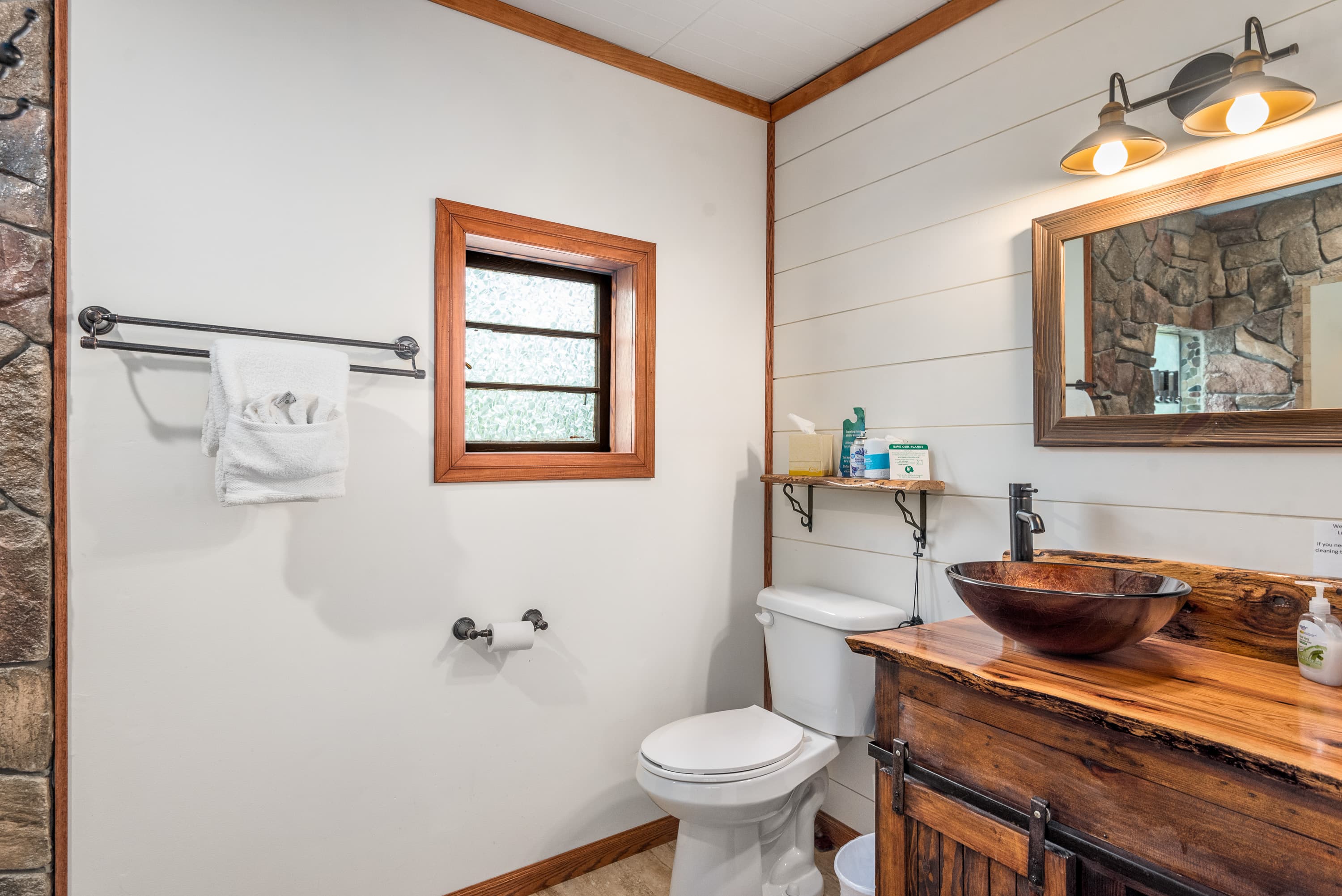 A cozy, rustic bathroom with a rock shower, wood plank walls, and a wooden vanity with a glass vessel sink. The room also features a toilet and a small window.
