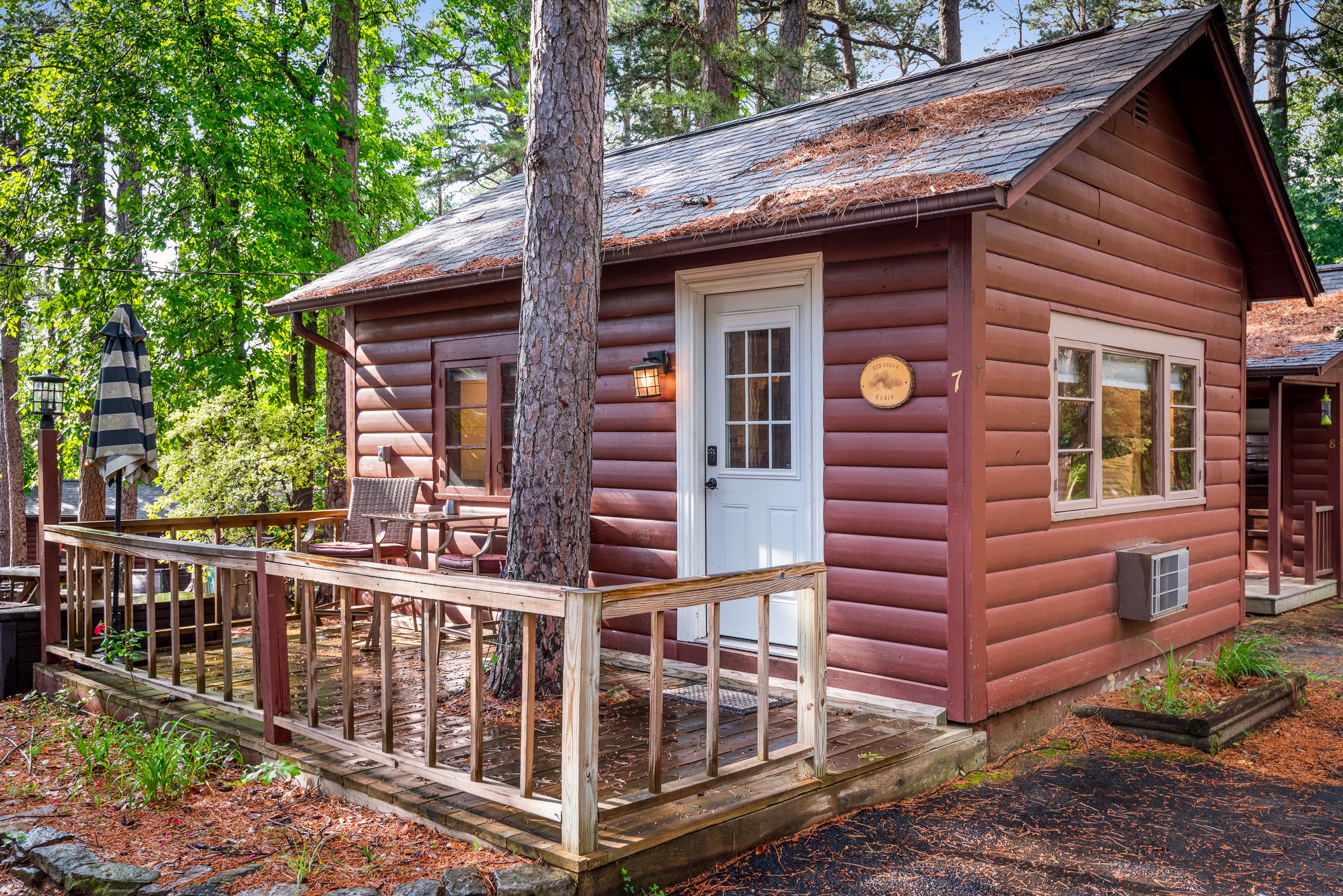 A rustic cabin is nestled among trees, with a small porch and a wooden deck. The deck features a table, two chairs, and a black patio umbrella.
