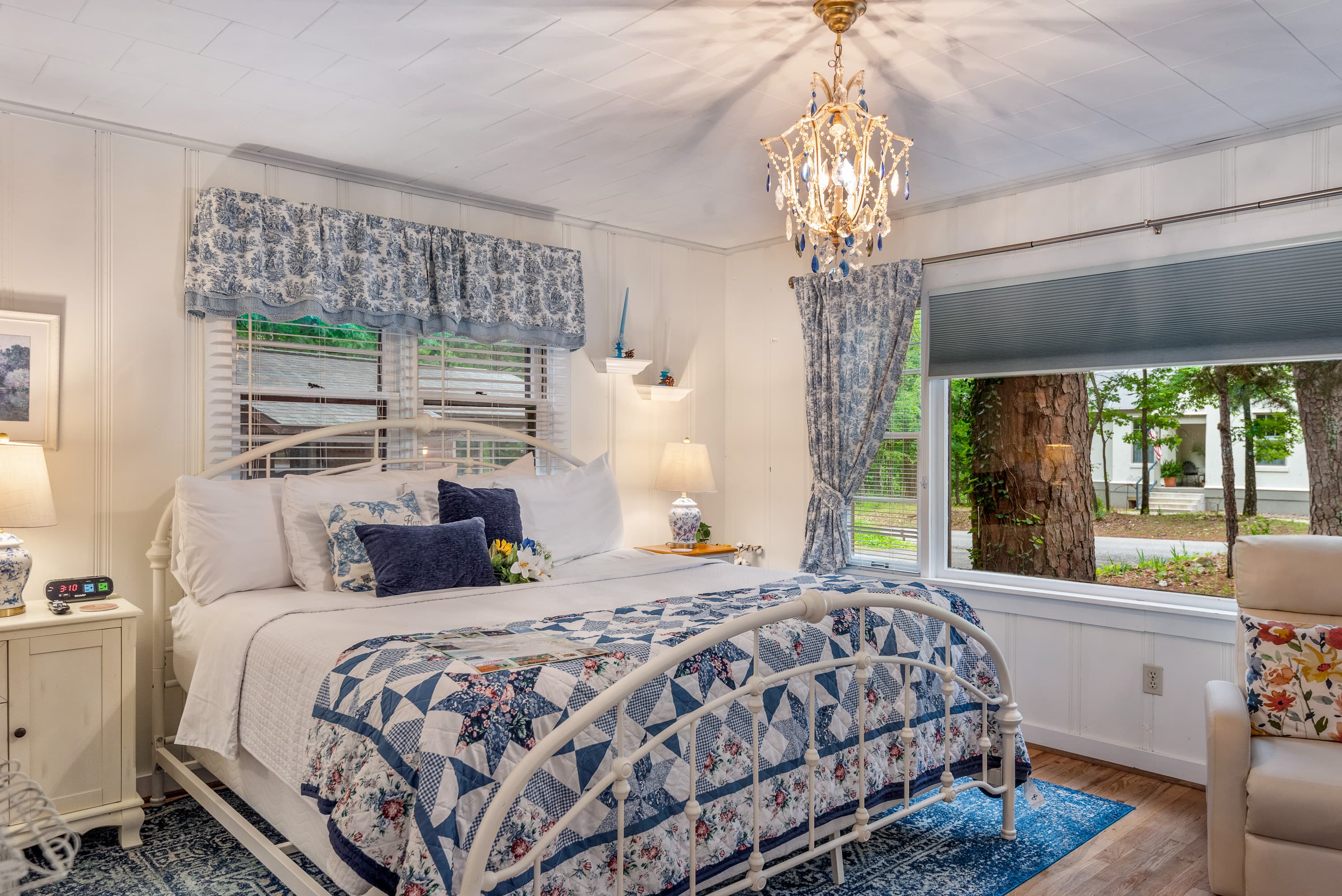 A king-size bed with a white metal frame and blue-and-white bedding is in a spacious bedroom. The room features a crystal chandelier and light-colored walls.