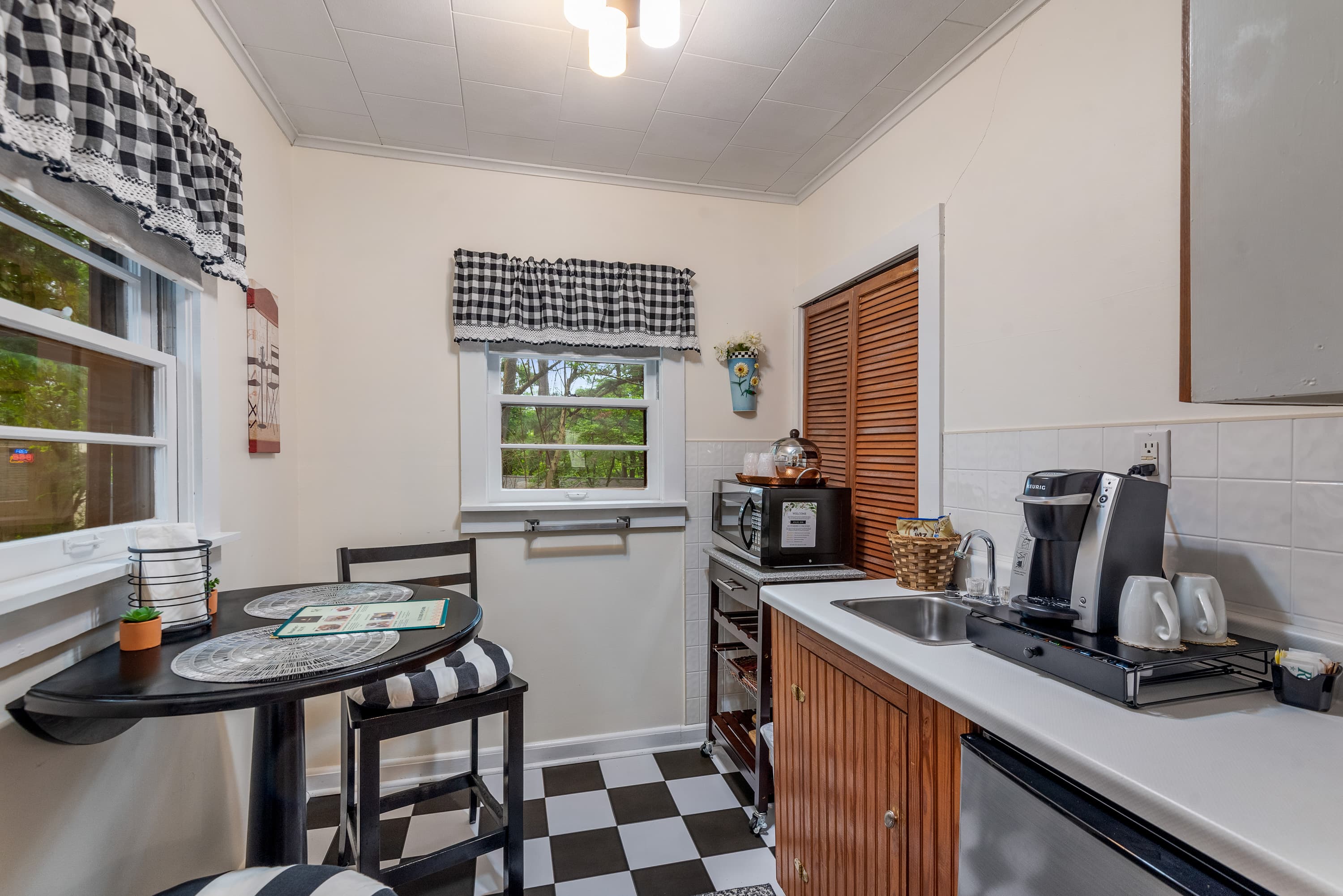 A compact kitchen features black and white checkerboard floors, a small table with two chairs, and a kitchenette area with a mini-fridge, microwave, and coffee maker.