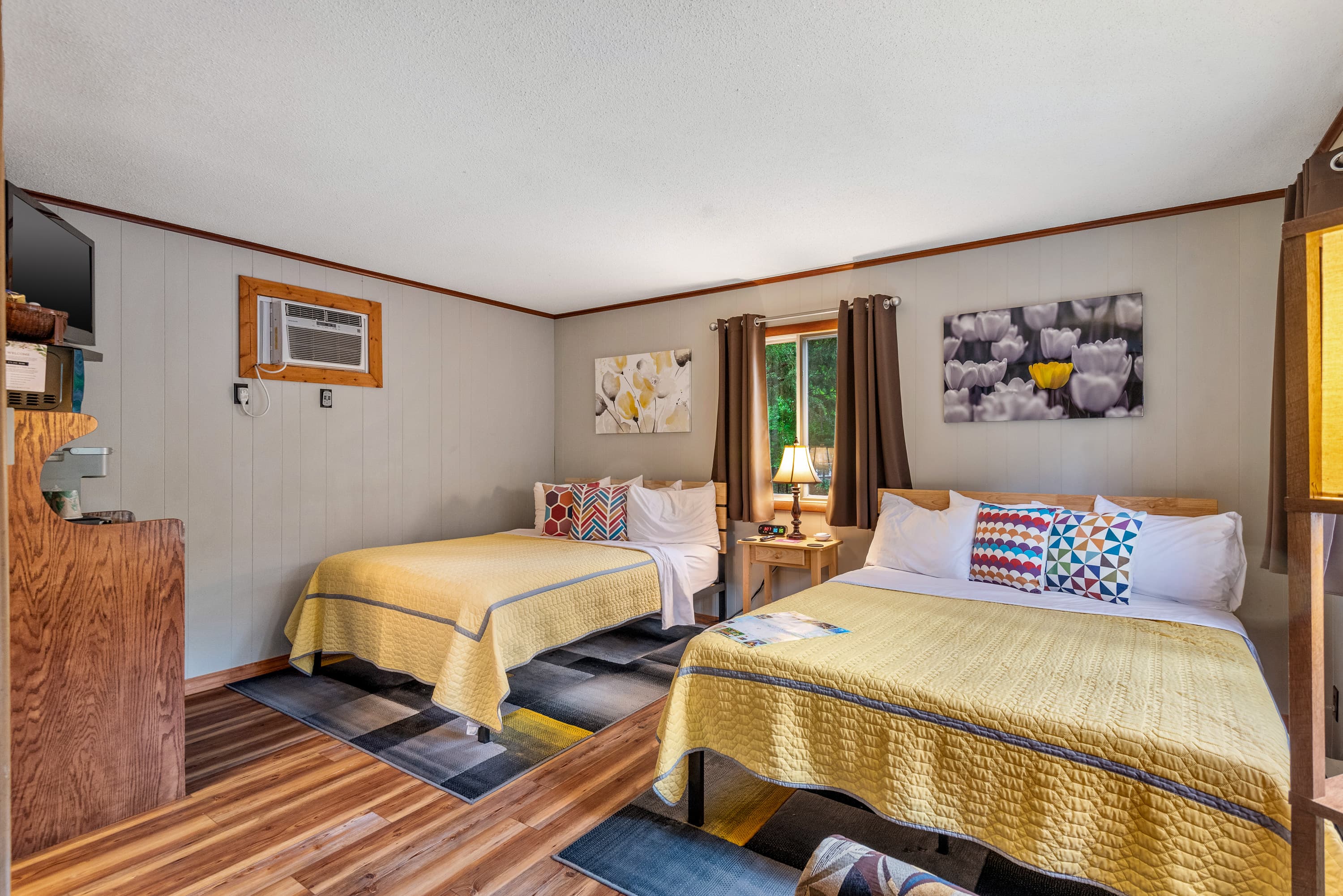 A rustic bedroom features two queen beds with yellow quilts and colorful pillows. The room has neutral-colored walls and a TV mounted on a wooden stand.