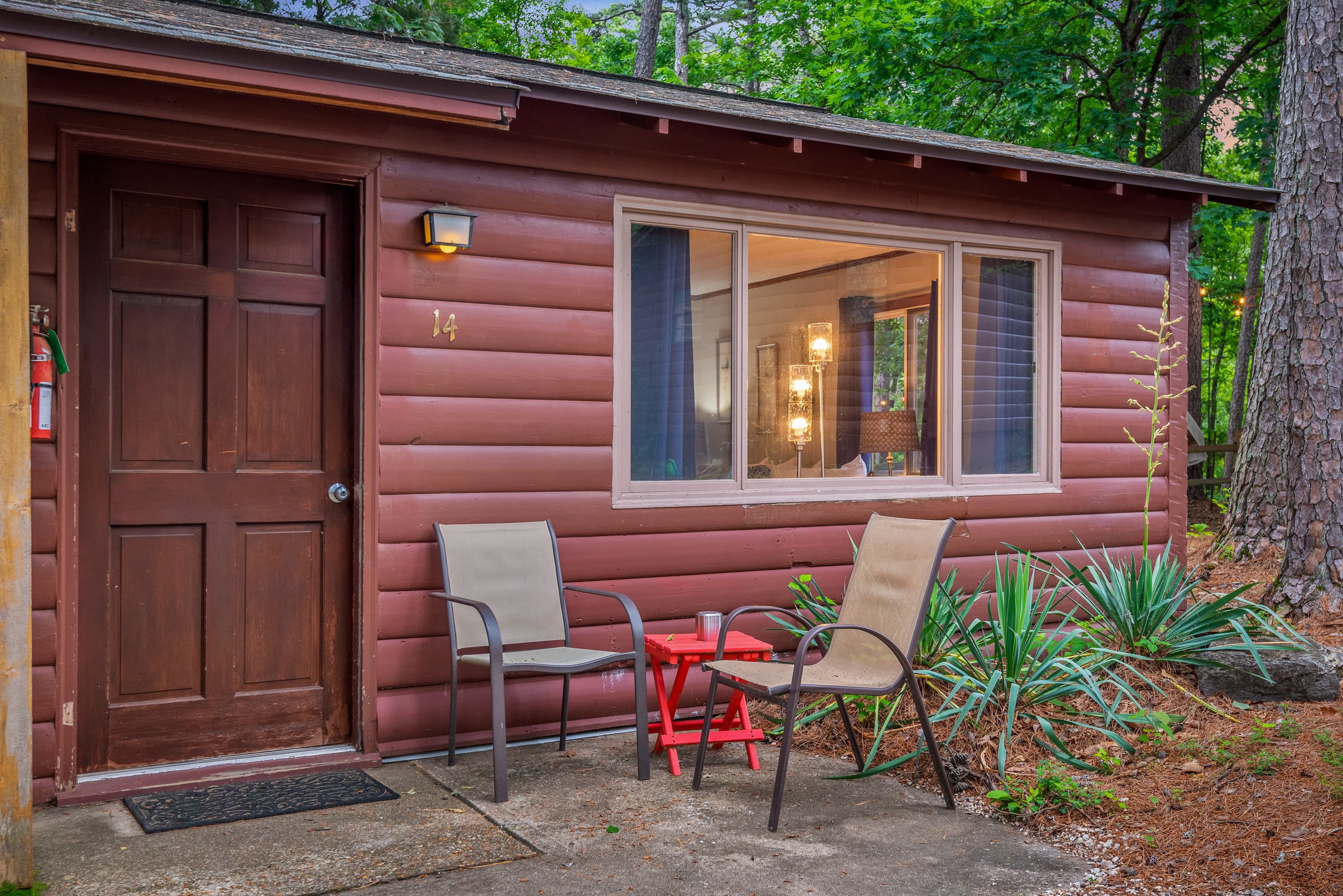 A rustic, red-brown duplex cabin with a paved driveway. Each unit has a small porch with a table and chairs, and there are windows with flower boxes.