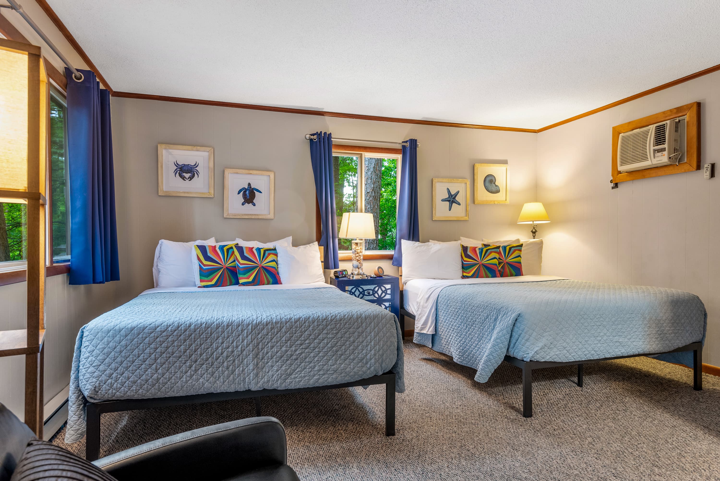 A rustic bedroom features two queen beds with quilted comforters and patterned pillows. The room has framed art of marine life and a nightstand between the beds.