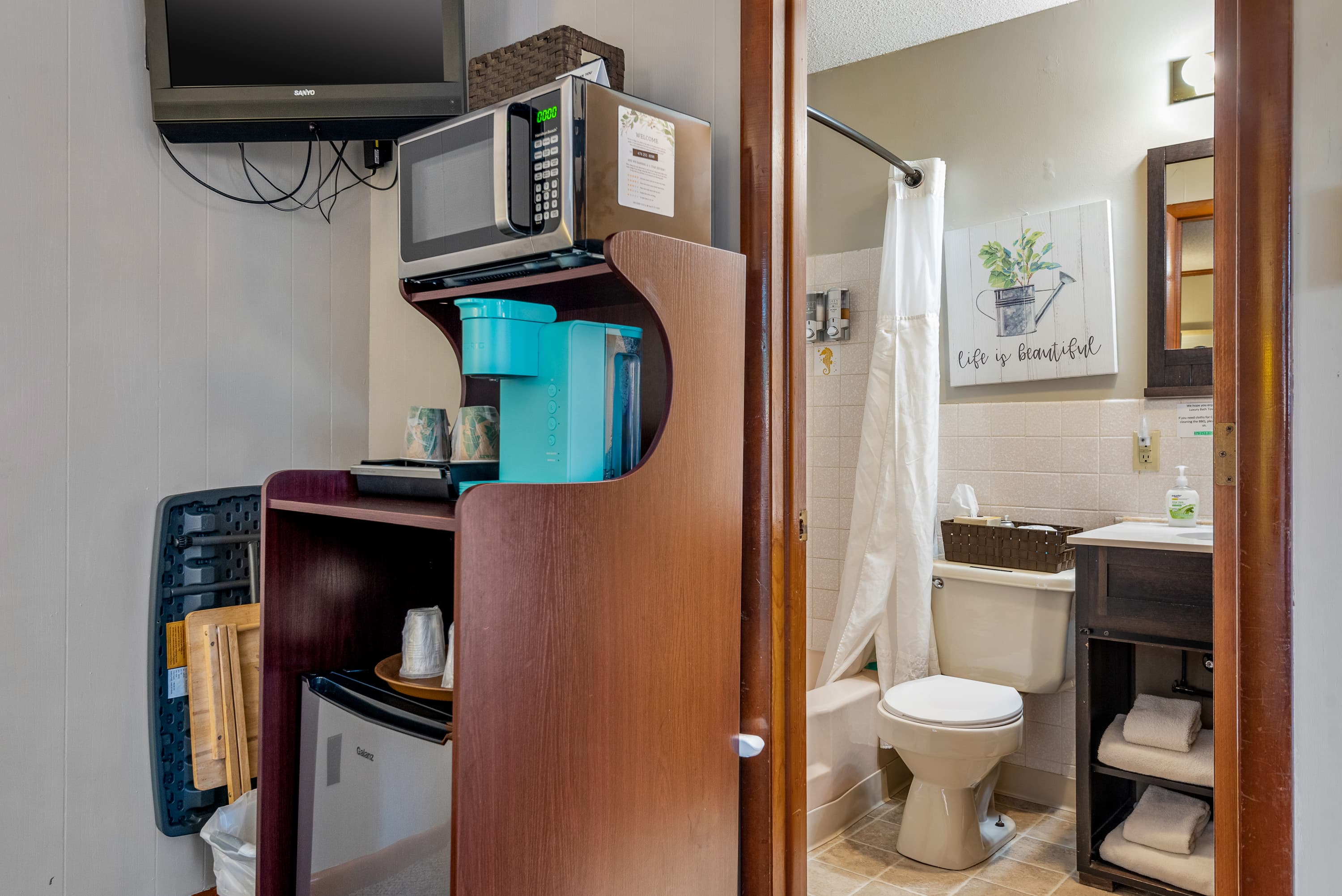 A clean, compact bathroom with a wood vanity and sink, a toilet, a shower with a white curtain, and a wall rack with neatly folded towels.
