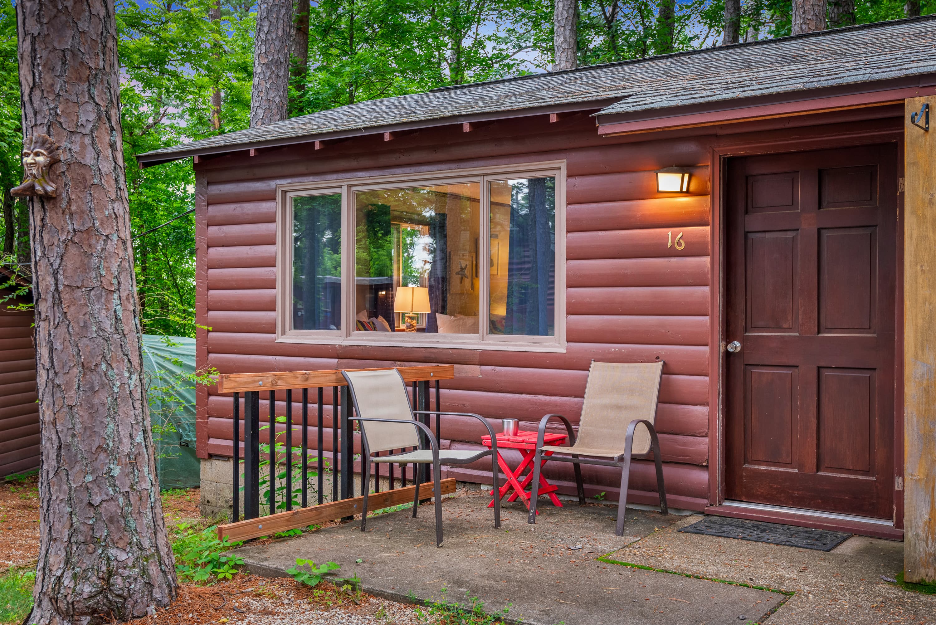 A rustic, red-brown duplex cabin with a paved driveway. Each unit has a small porch with a table and chairs, and there are windows with flower boxes.