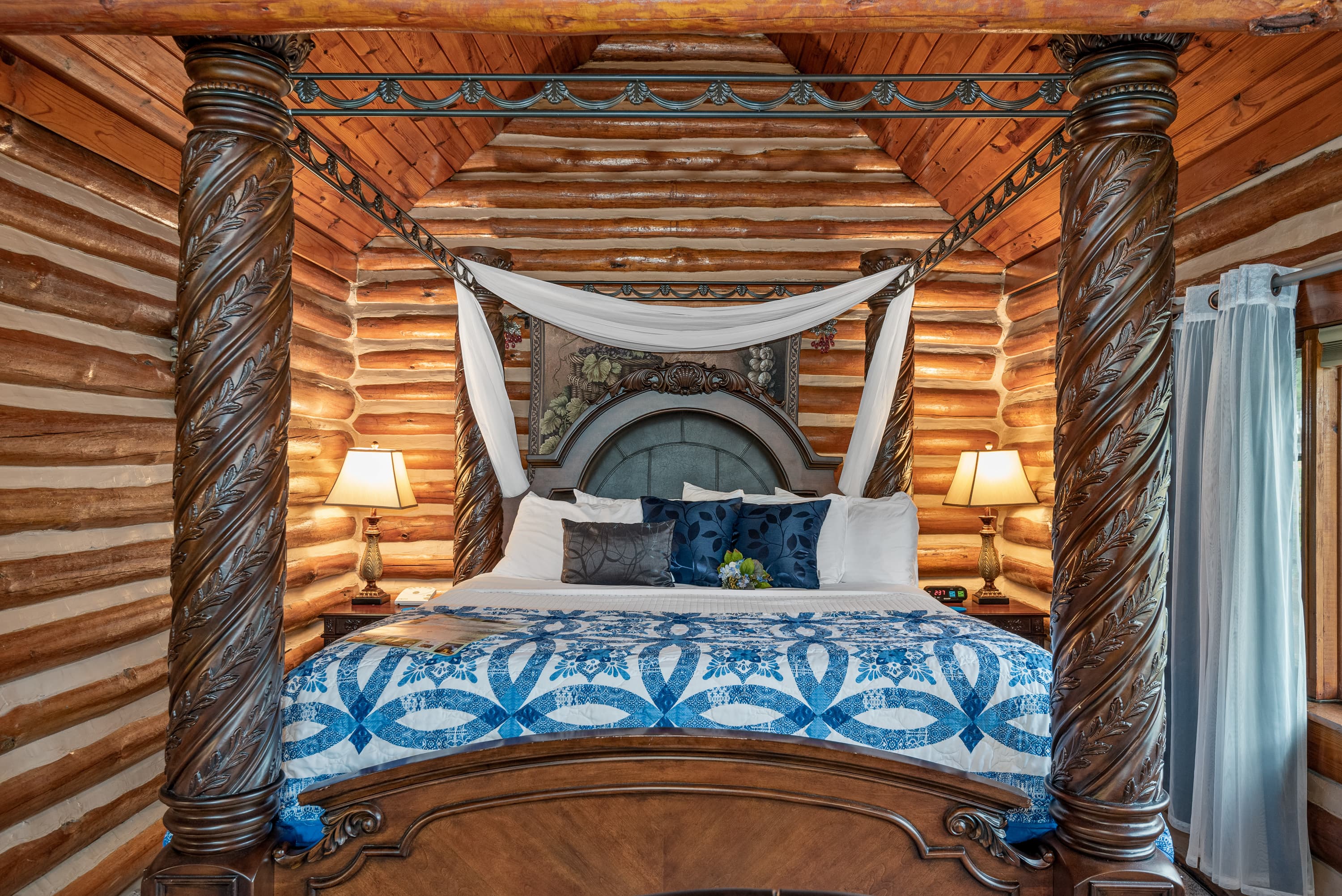 A cozy, rustic bedroom with a stunning king-size, four-poster bed. The room features wood log walls, a beautiful bed quilt, and two bedside lamps.