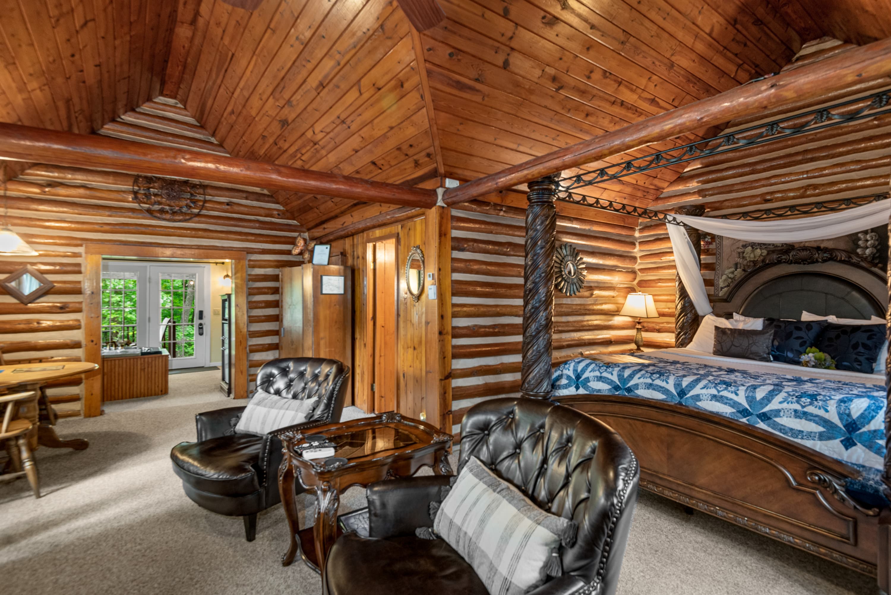 A spacious, rustic log cabin bedroom with a king-size, four-poster bed, two leather armchairs, and a small table. The room has high vaulted wood ceilings and log walls.