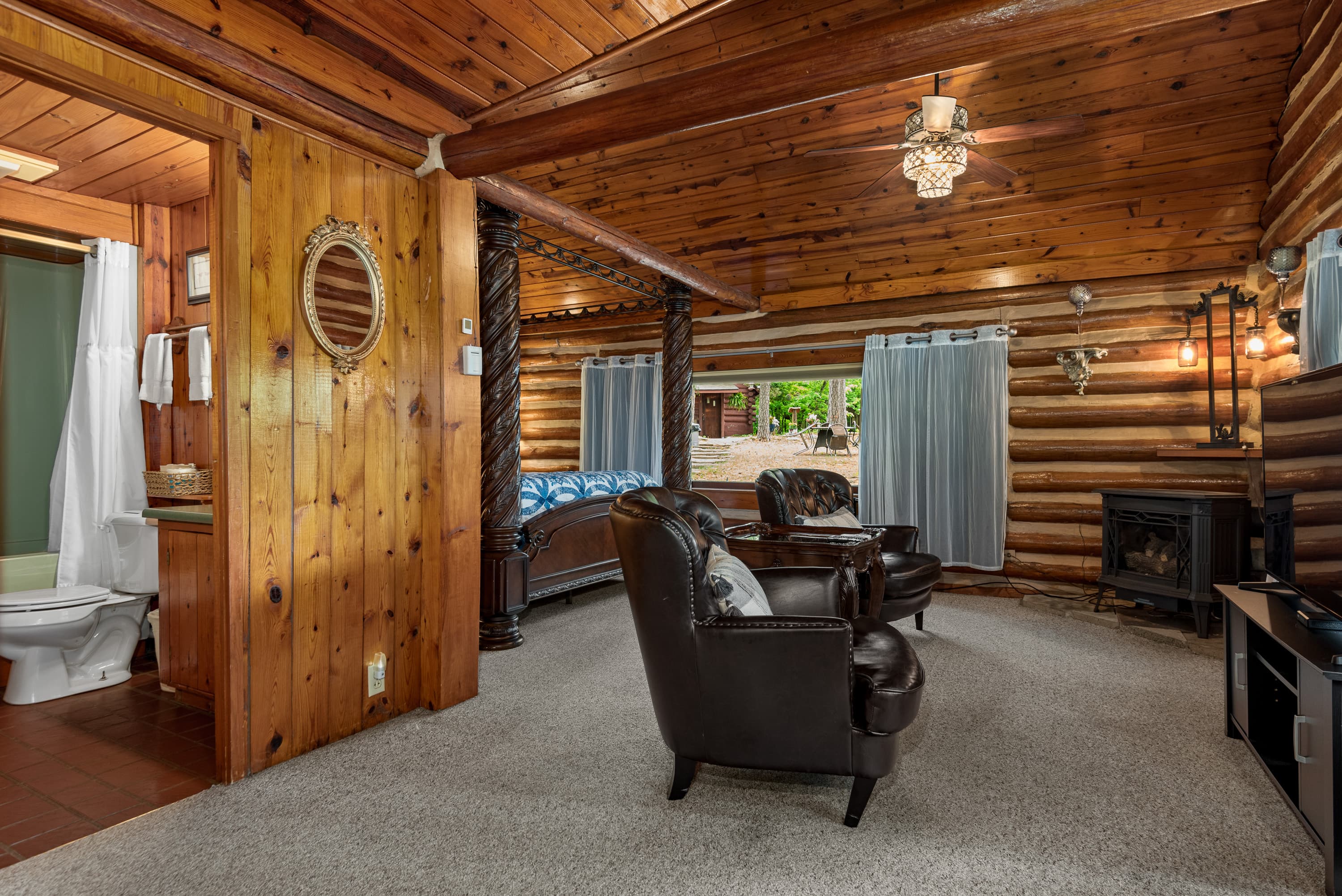 A spacious, rustic log cabin studio with a king-size bed, two leather armchairs, and a fireplace. An open doorway leads to a small bathroom.