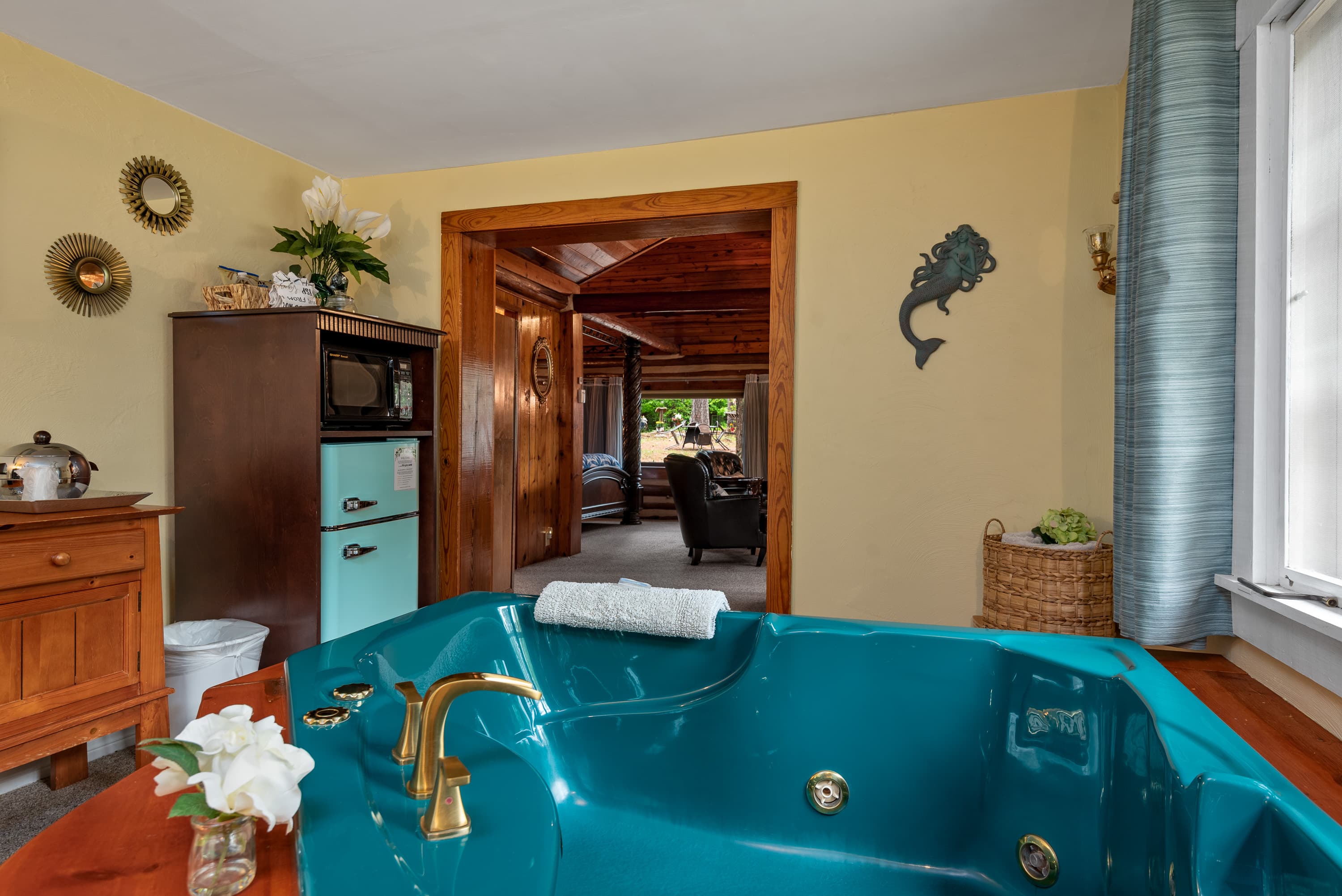 A sunroom with a turquoise jetted tub and a mini-fridge. A doorway leads to a living area with two leather armchairs and a small table.
