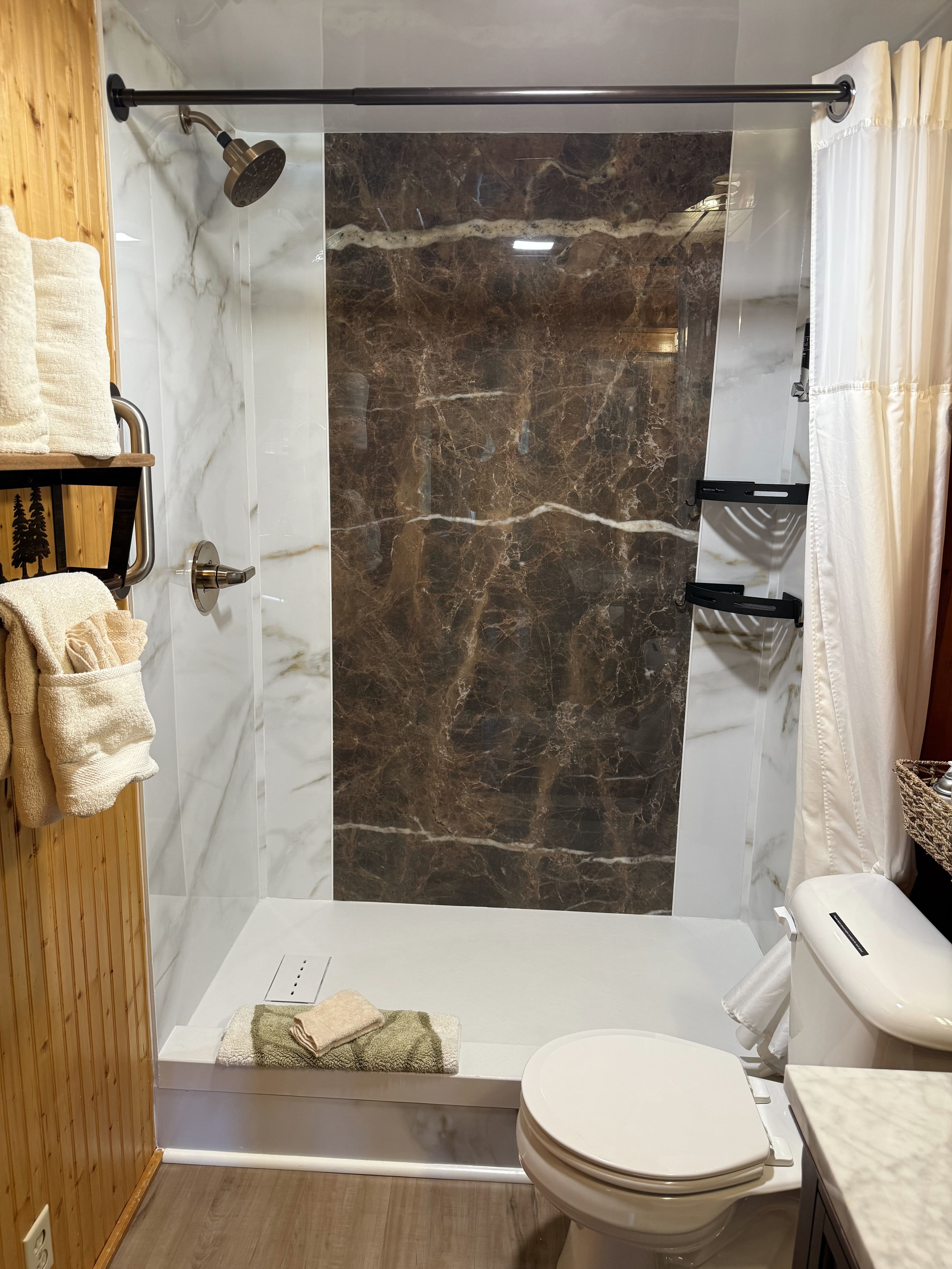 Shower featuring brown and white marble walls, a black showerhead, towel rack with towels, a toilet, and a white curtain.