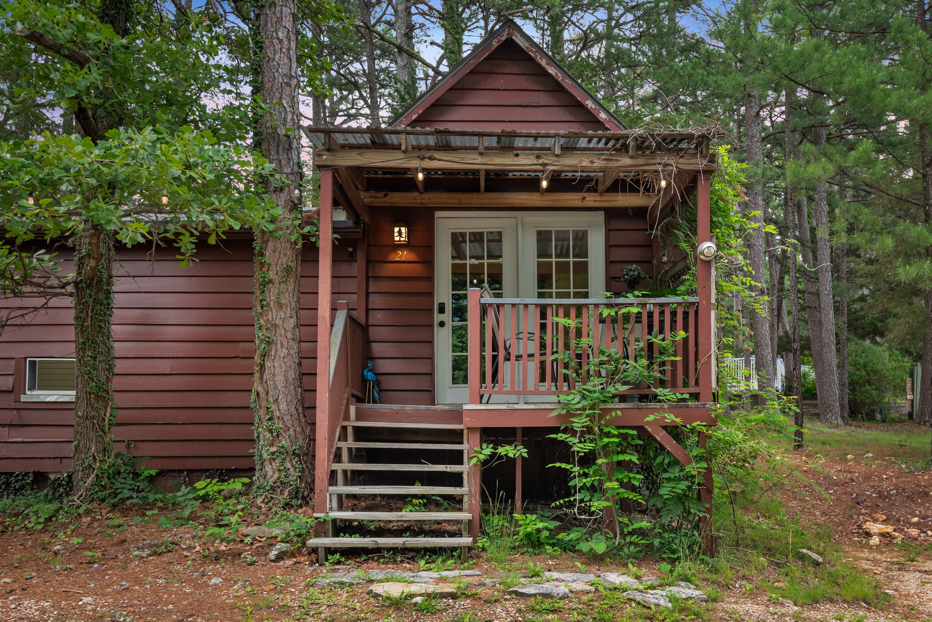 A rustic, wood-paneled cabin with a small porch and a deck. A few steps lead to a front door with a window. The cabin is surrounded by trees.