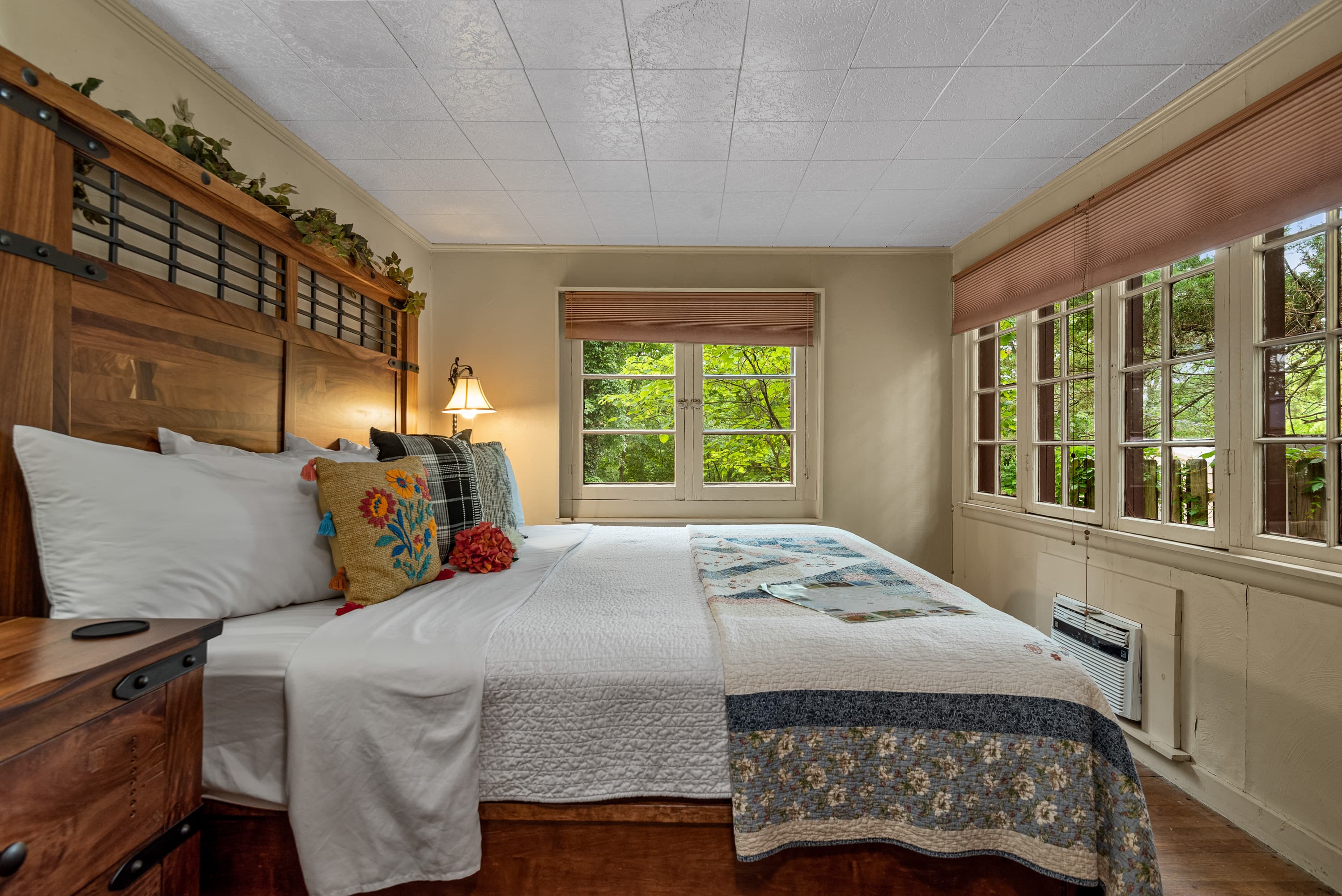 A spacious bedroom features a king-size bed with a white comforter and blue floral quilt. The room has wood floors, a paneled ceiling, and large windows.