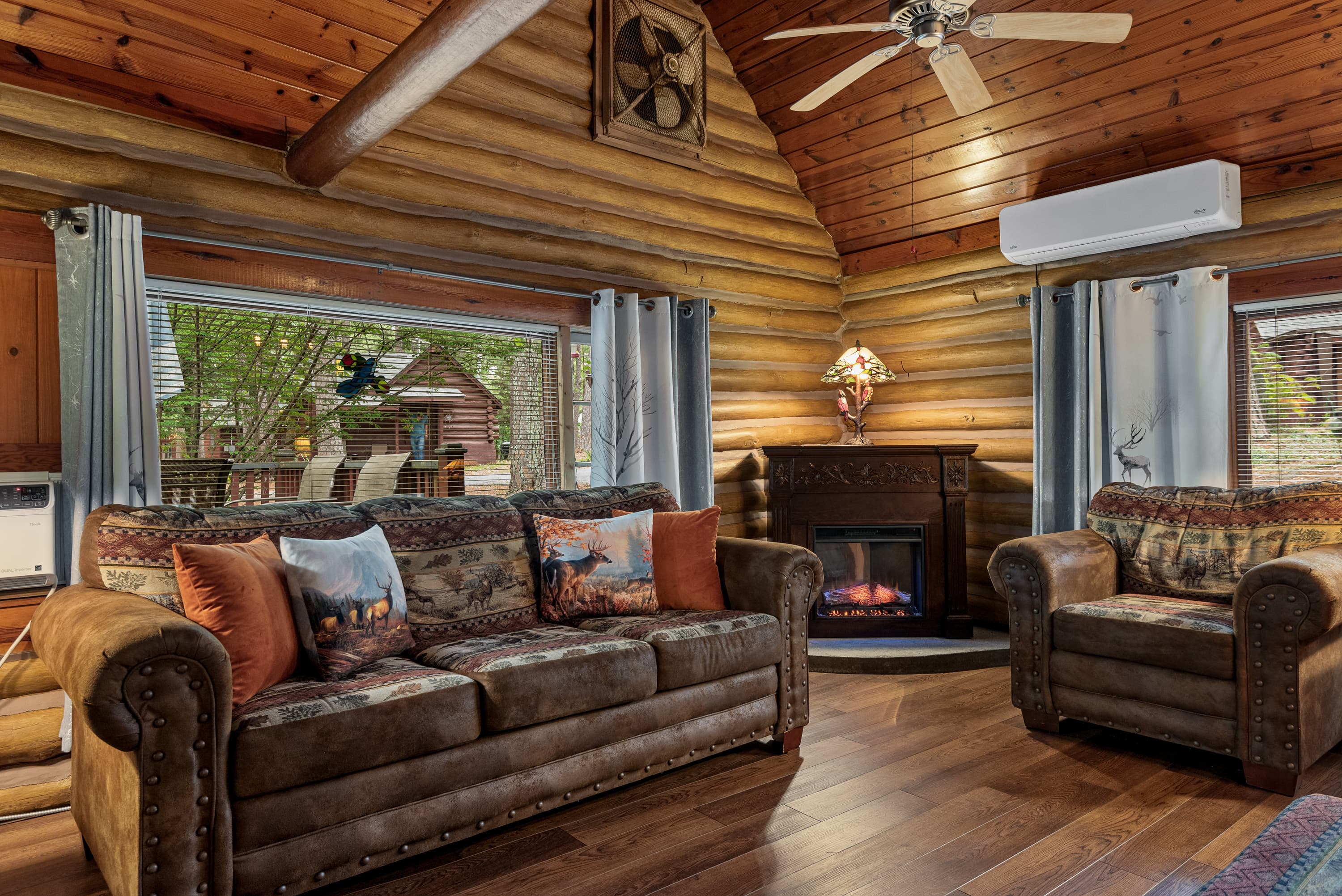 A cozy log cabin living room features a brown leather couch, an armchair, and a fireplace in the corner. The room has high vaulted ceilings and a ceiling fan.