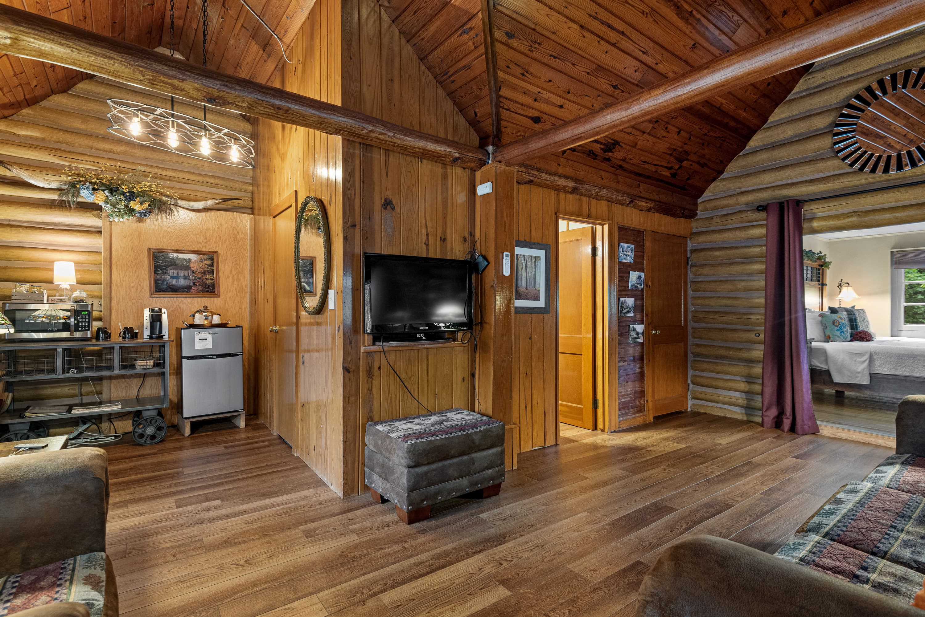 A cozy, open-concept log cabin living room with a wood-paneled ceiling and hardwood floors. The room features a flat-screen TV mounted on a small dividing wall.