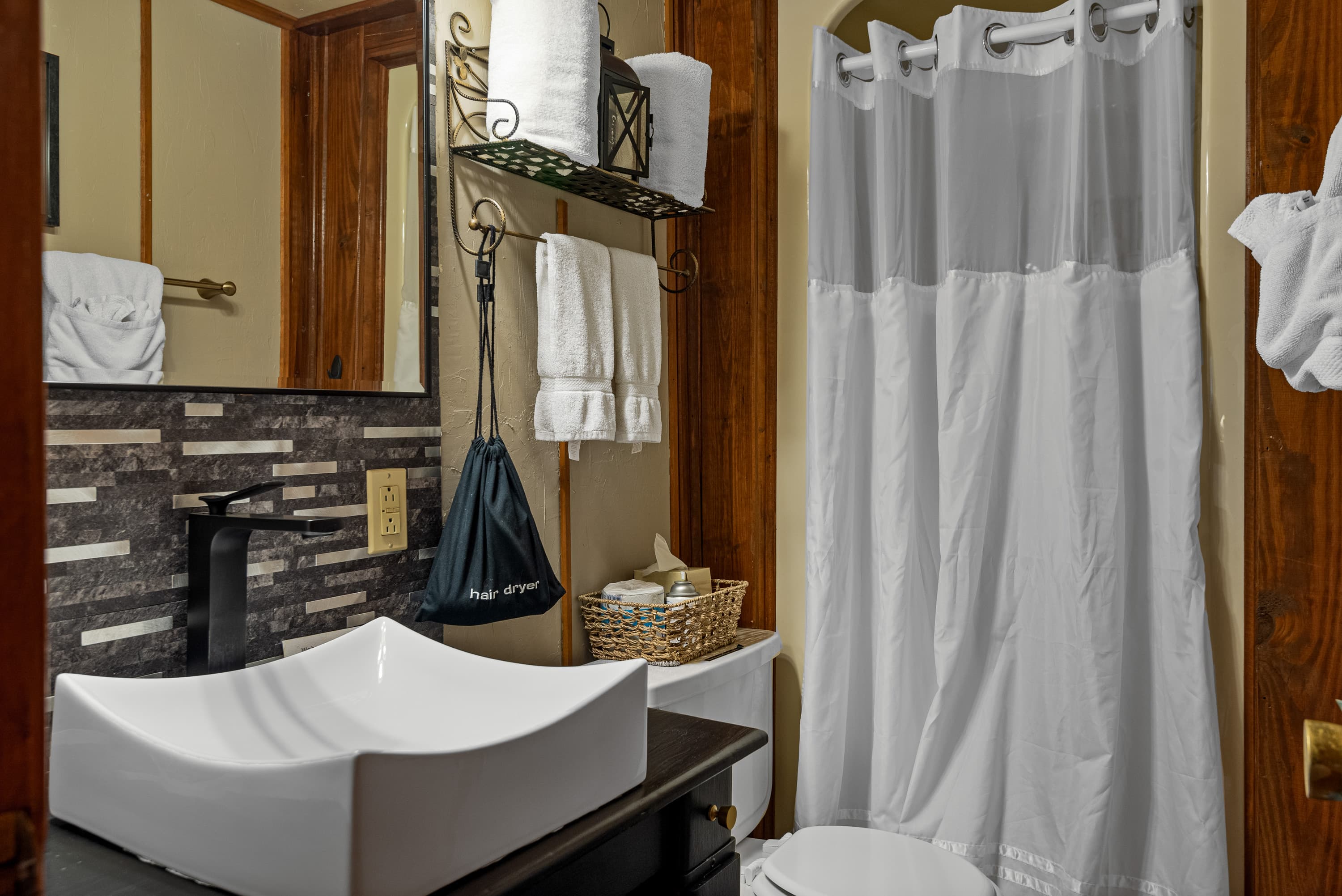 A small, rustic bathroom with a black vanity and a white vessel sink. The room also features a toilet, a shower with a white curtain, and a mosaic tile backsplash.