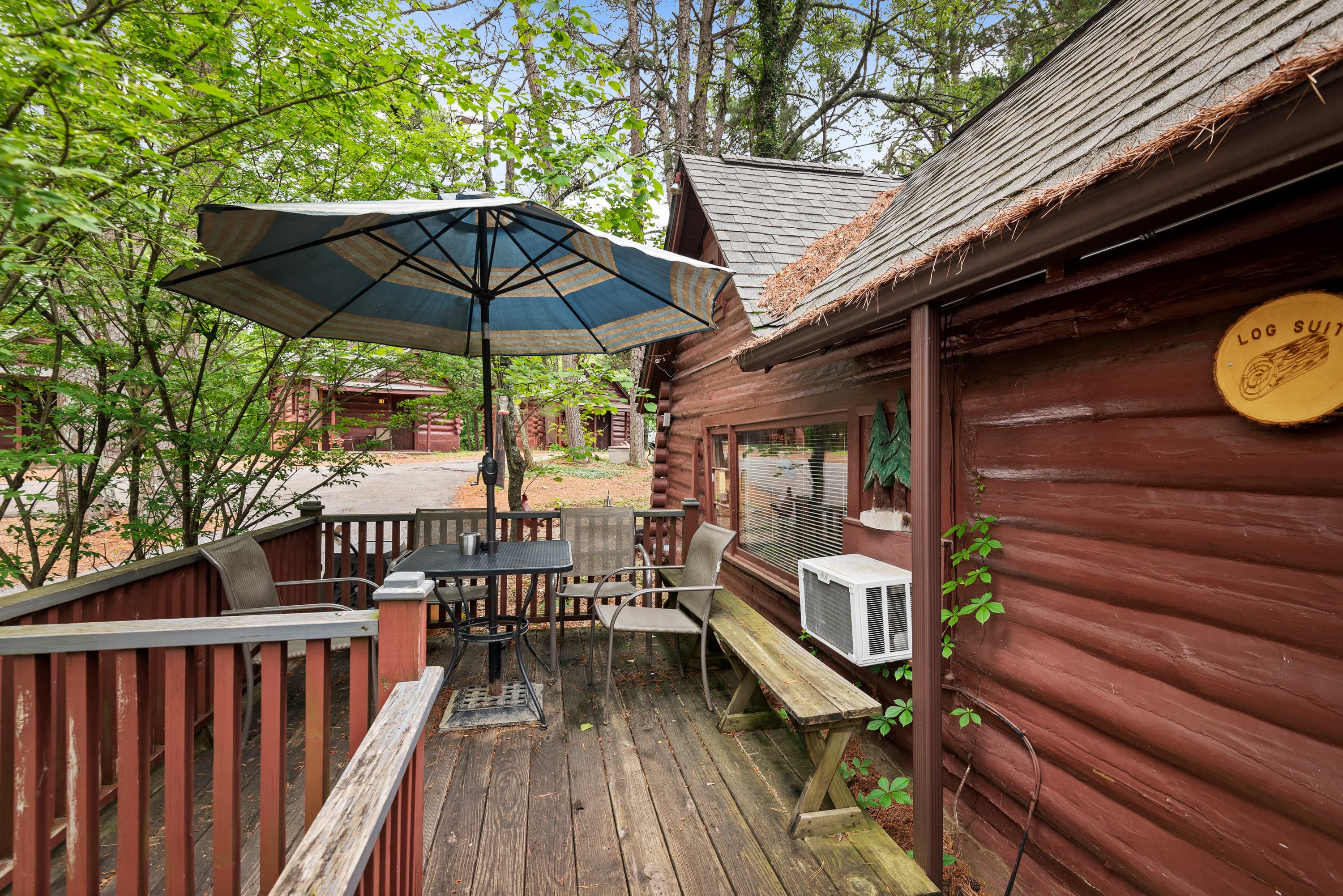 A small, rustic cabin with a front porch and a deck. The deck features a round table, chairs, a bench, and a large umbrella.