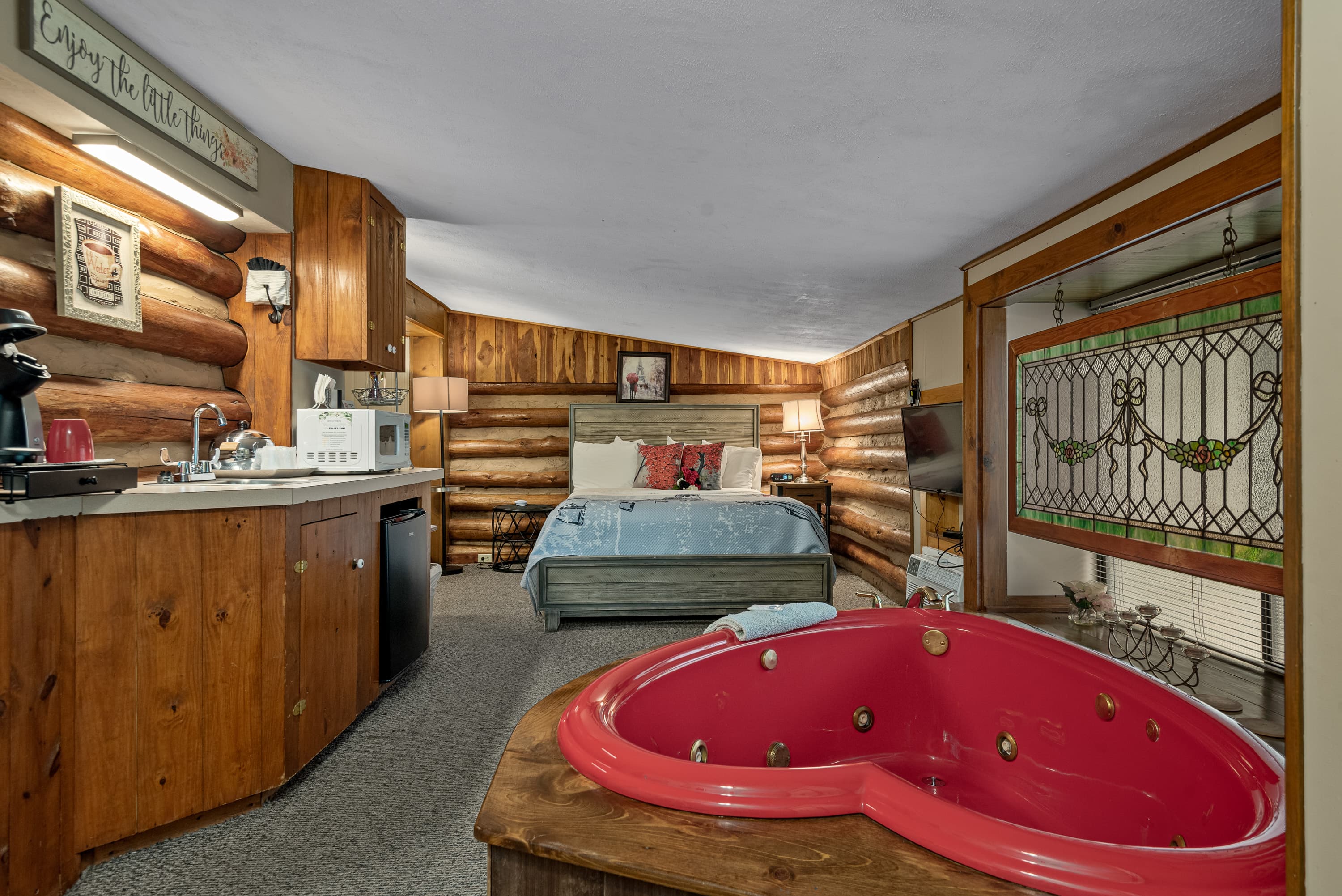 A log cabin bedroom features a red heart-shaped jetted tub, a king-size bed, a small kitchenette with a mini-fridge and a coffee maker, and rustic wood log walls.