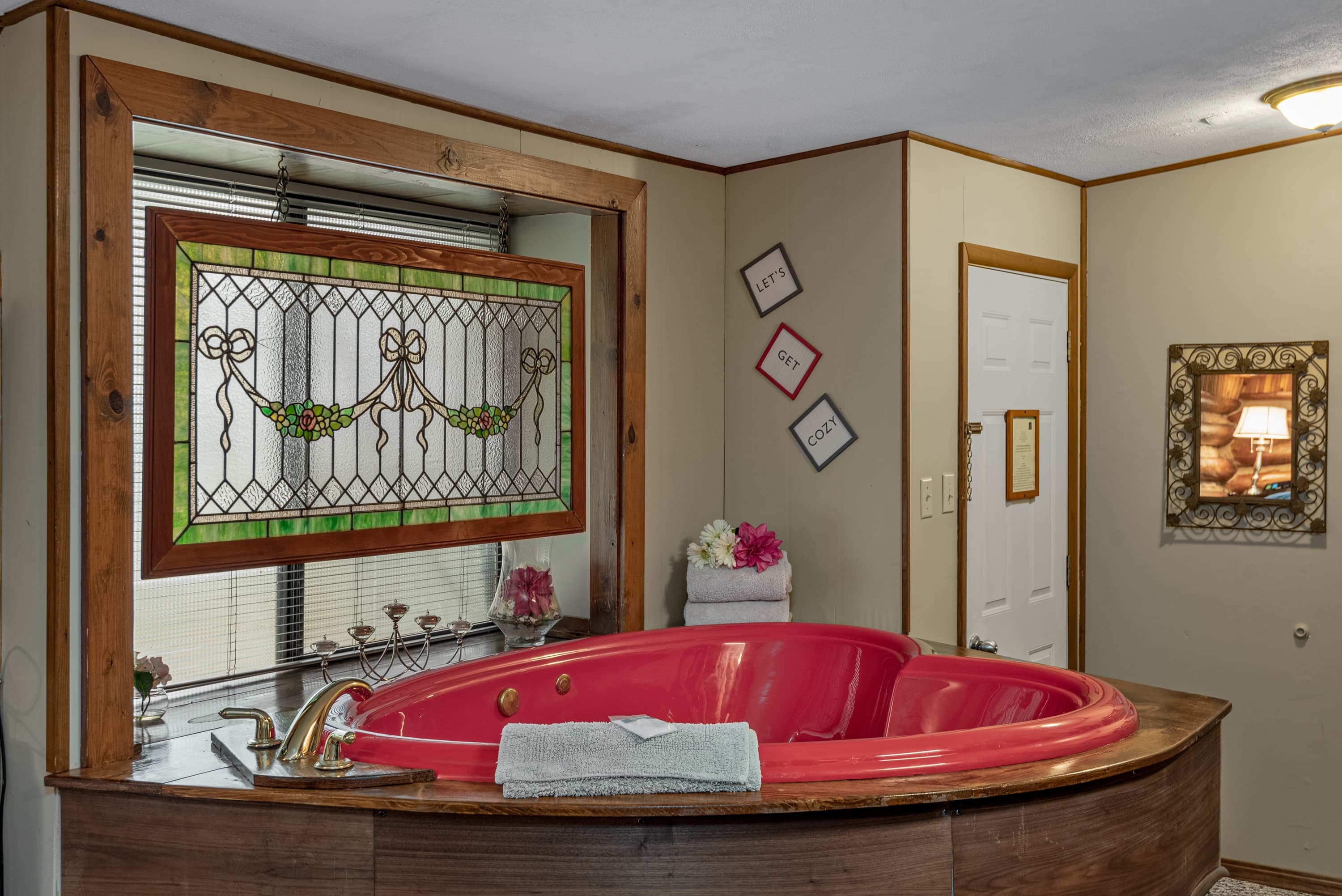 A rustic bedroom with a red, heart-shaped jetted tub. The space has a stained-glass window and a wall with three small framed pictures.