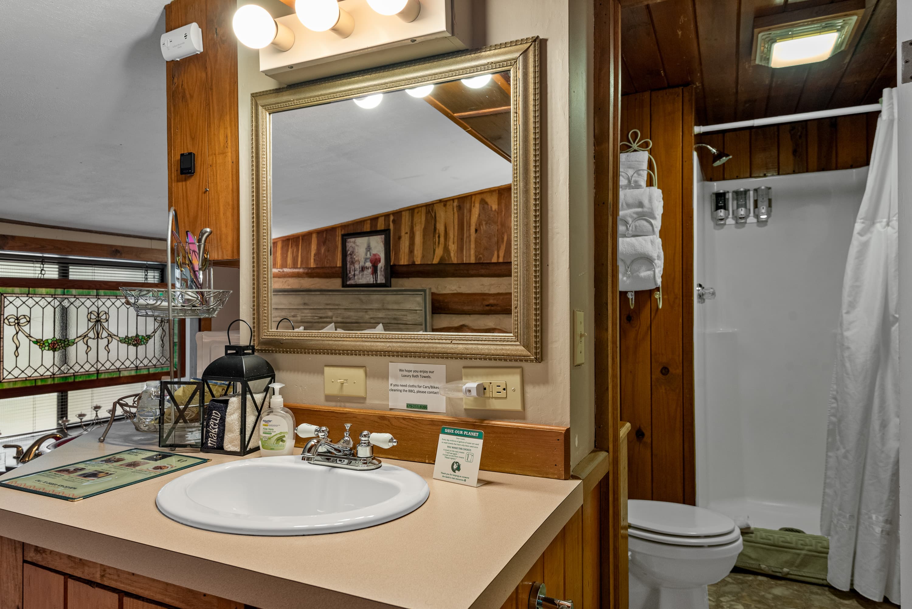 A small, rustic bathroom with a long counter and a white sink. The space has wood-paneled walls and a shower with a white curtain.
